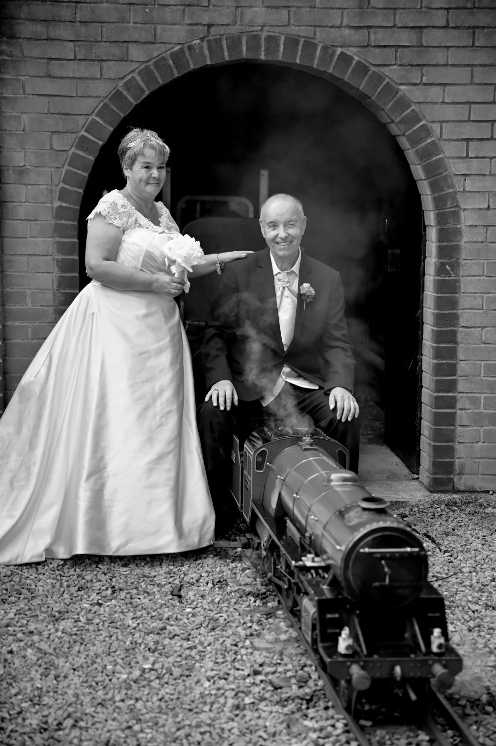 A bride and groom in wedding attire sitting on a miniature train outside a brick tunnel, with a cloud of smoke rising from the locomotive, captured in black and white.