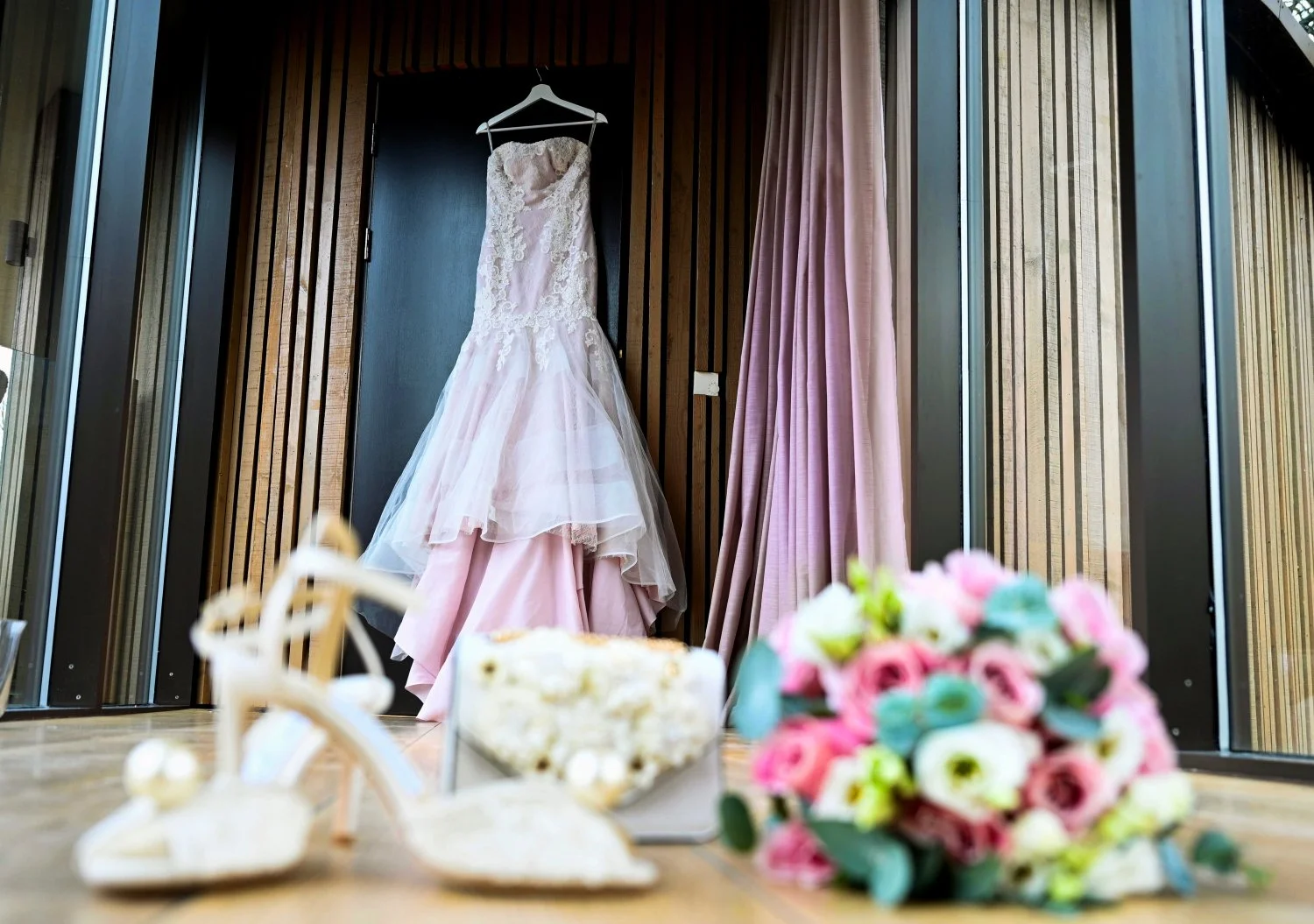 Blush pink lace wedding dress hanging in luxury treehouse bridal suite, heels, pearl clutch bag and pink rose bouquet in foreground, wedding detail photography Chewton Glen New Forest Hampshire