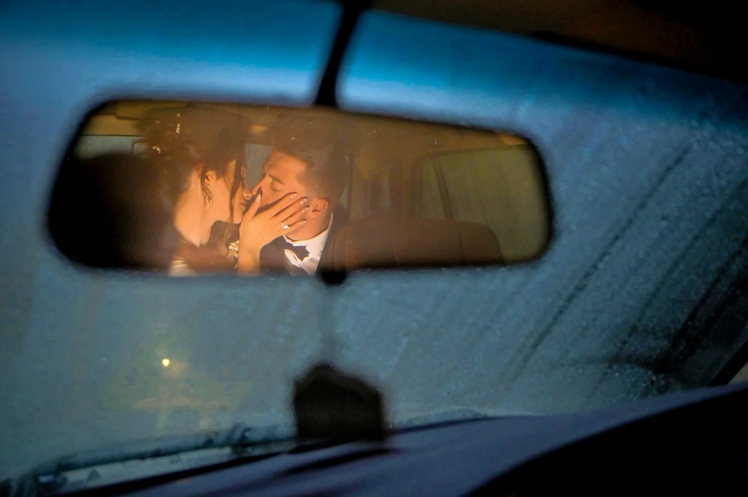 A couple in formal attire sharing an intimate moment inside a vehicle, seen through the rearview mirror. The woman is touching the man's face, and they are about to kiss.