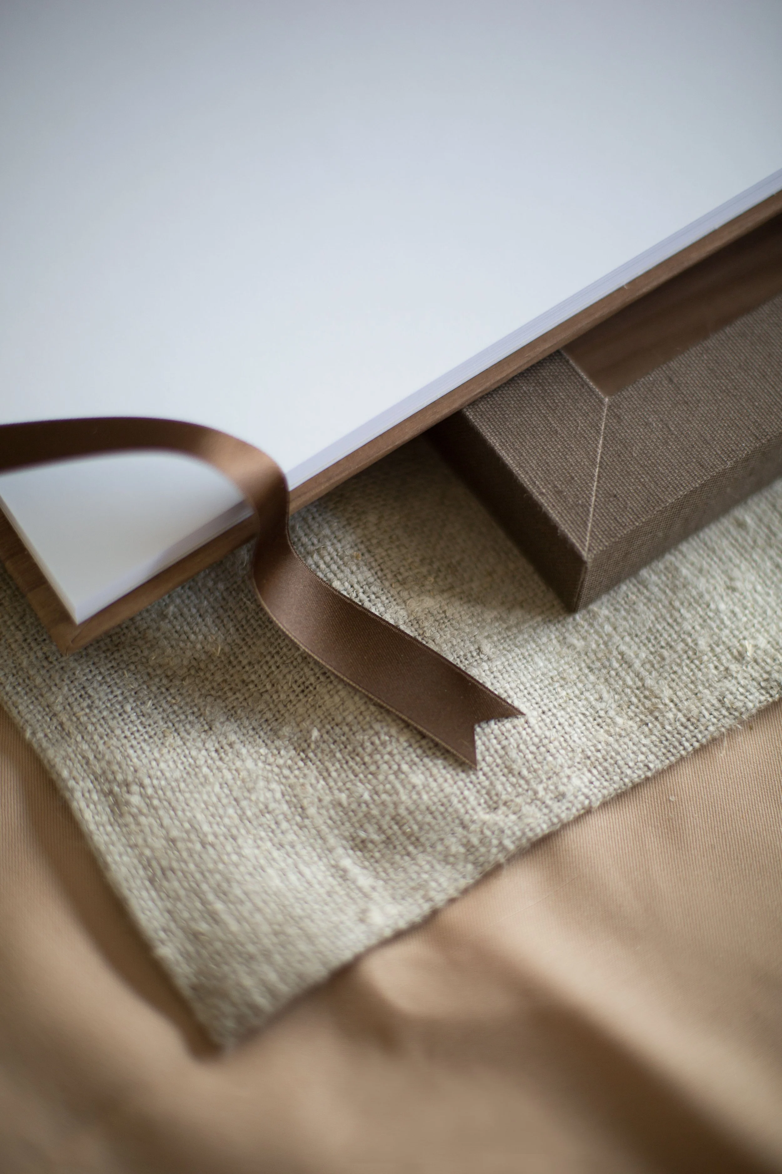 Close-up of a notebook or album with white pages, bound with a wooden or wood-like cover, placed on a beige fabric surface with a textured look.