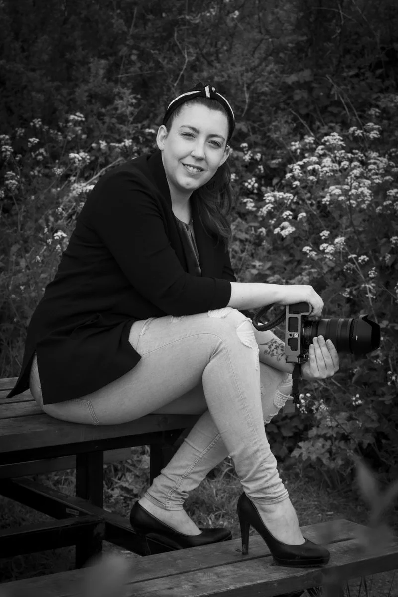 A woman sitting on a wooden bench outdoors, holding a camera with a large lens, surrounded by bushes with small flowers in the background, black and white photo.