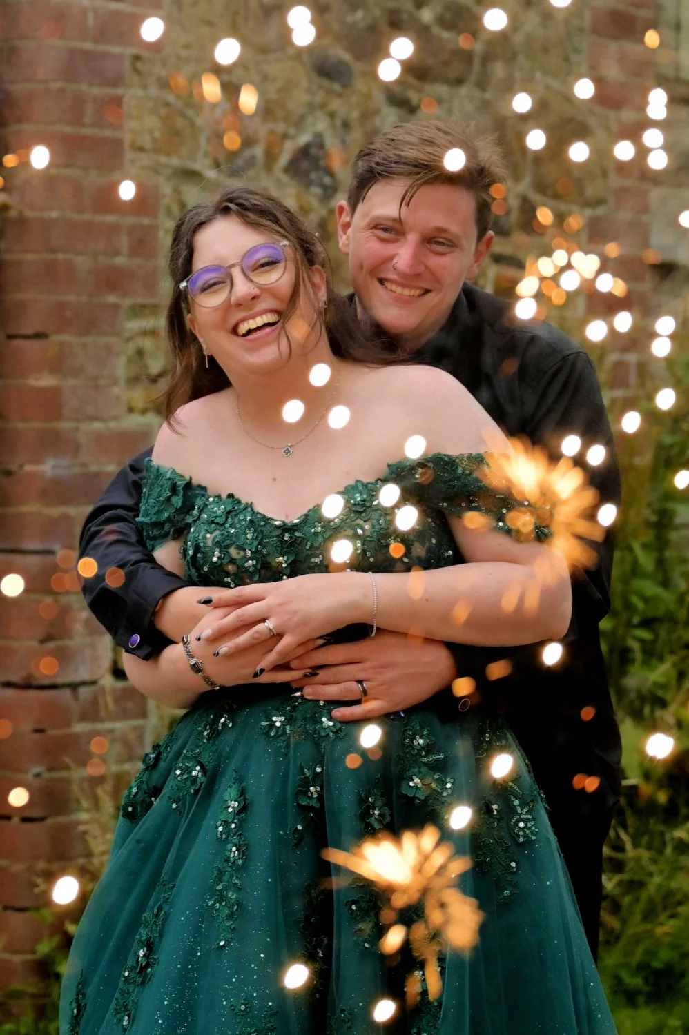 A smiling woman in a green dress with sequins and a man in black, celebrating outdoors with sparklers and string lights behind a brick wall.