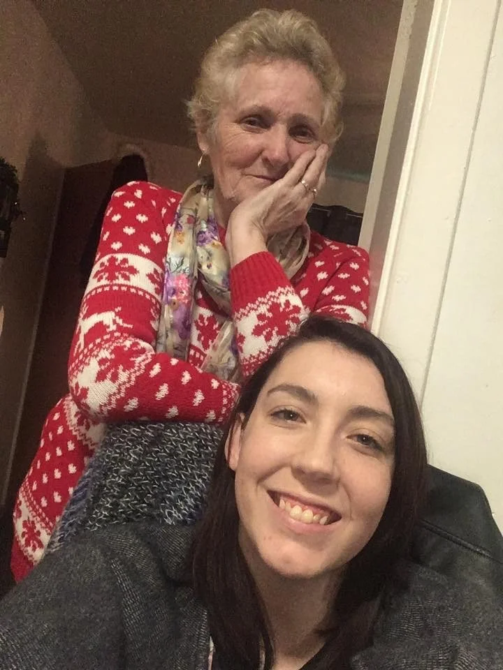 A young woman and an older woman smiling for a selfie indoors. The older woman is wearing a red sweater with white snowflakes, a colorful scarf, and has short, curly hair. She is resting her face on her hand with a gentle smile. The younger woman, sitting in front, has long dark hair, is wearing a gray blazer, and is smiling at the camera.