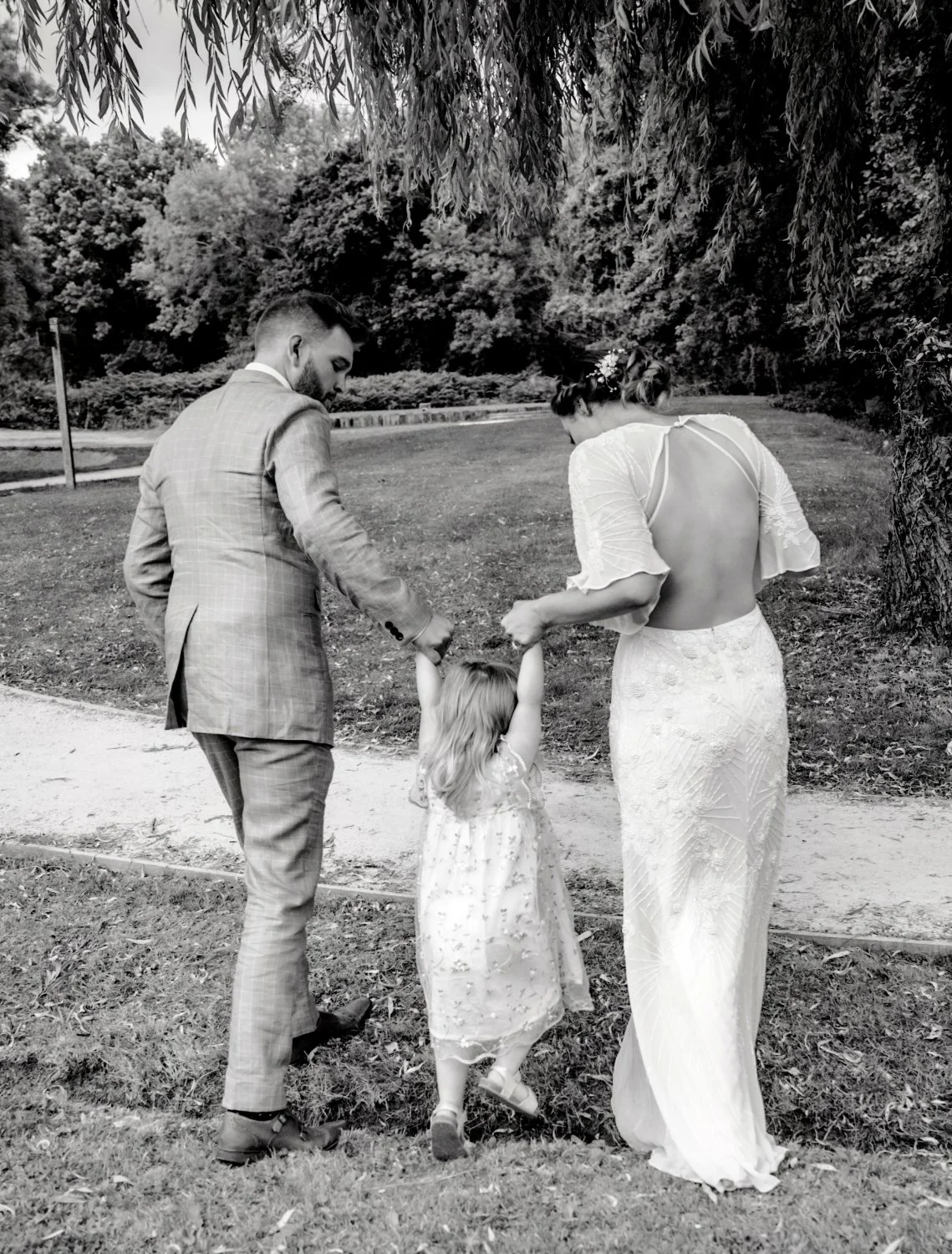 Black and white candid wedding portrait of bride and groom holding hands with young flower girl under weeping willow tree, documentary family wedding photography