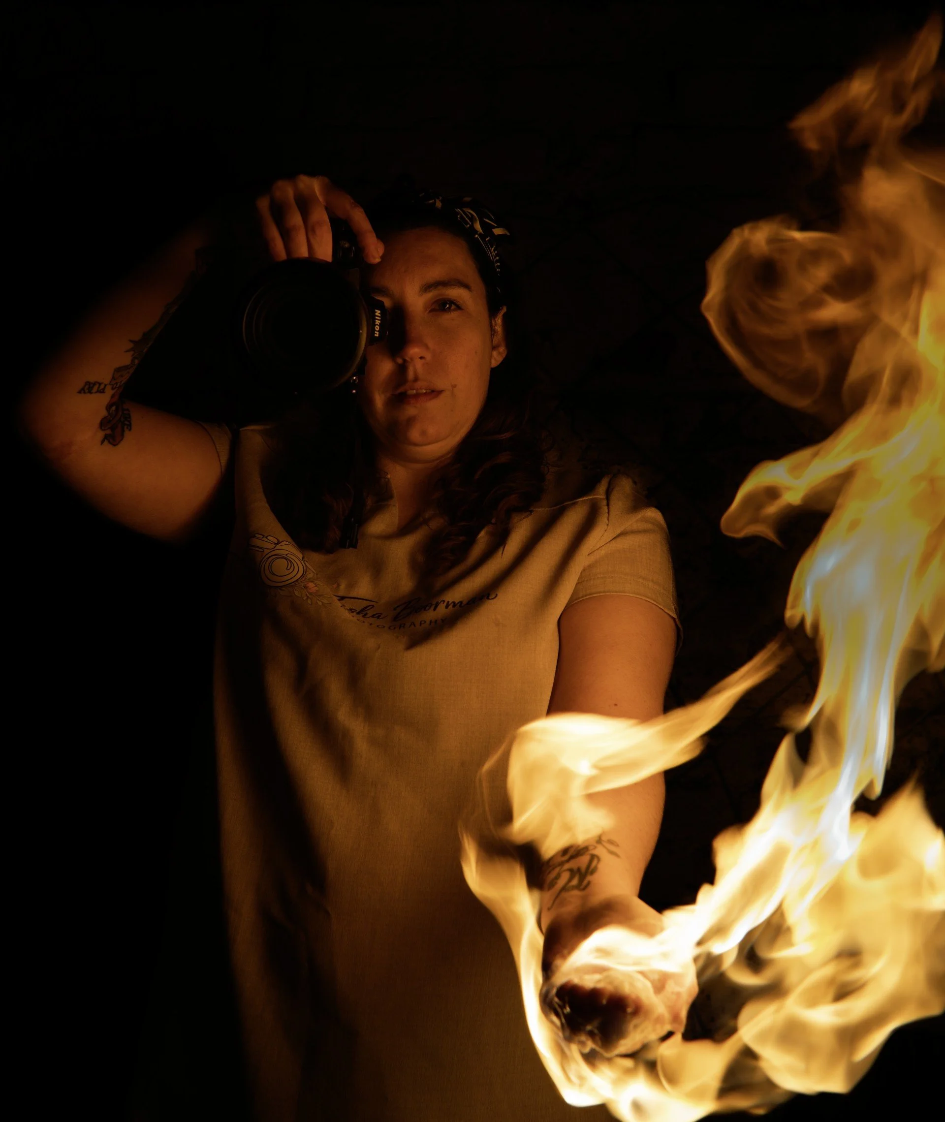A person with tattoos on their arm and a patterned headband is taking a mirror selfie with a DSLR camera. They are lying on the ground near a fire, which is creating bright flames and illuminating their face with warm light in a dark setting.