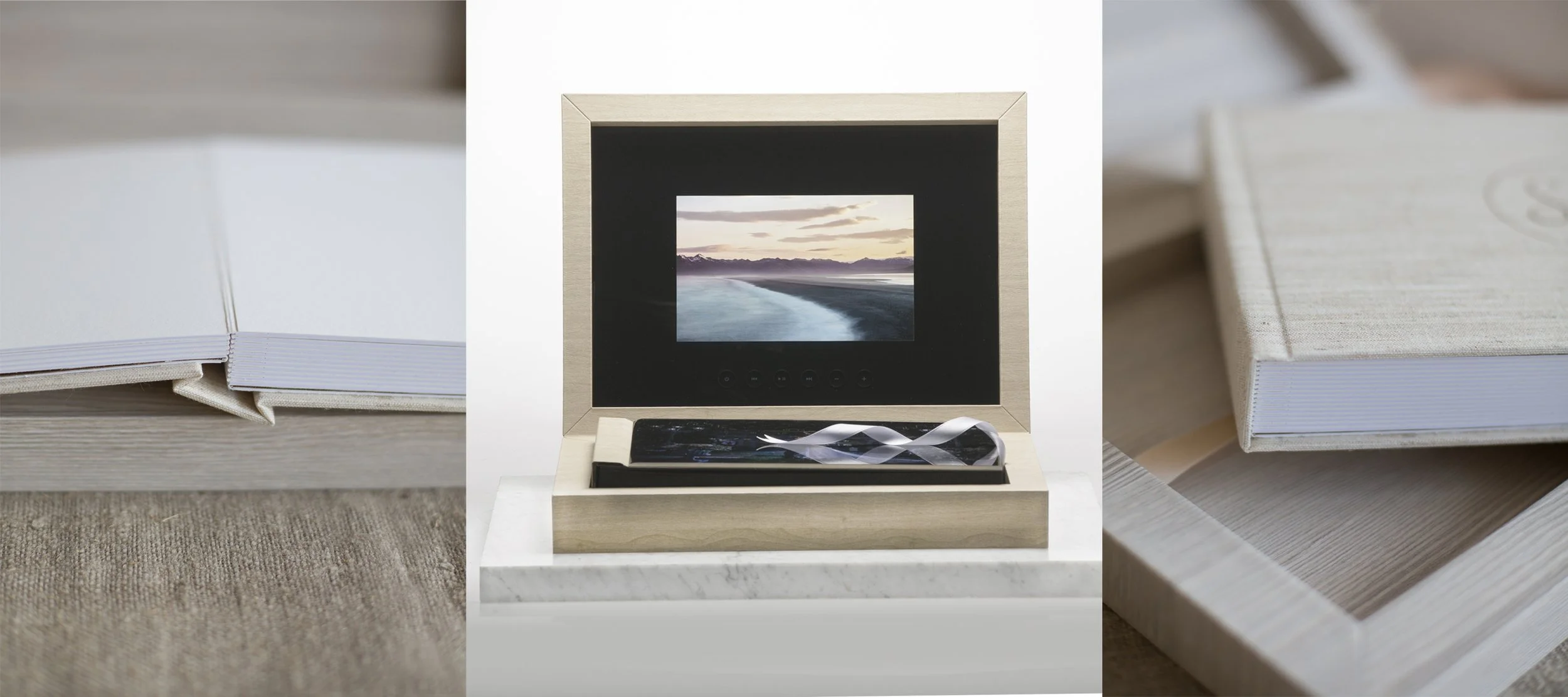 A framed photo of a mountain landscape with water, set in a beige box with control buttons, placed on a white marble surface, flanked by two beige books or albums on a light wooden table.