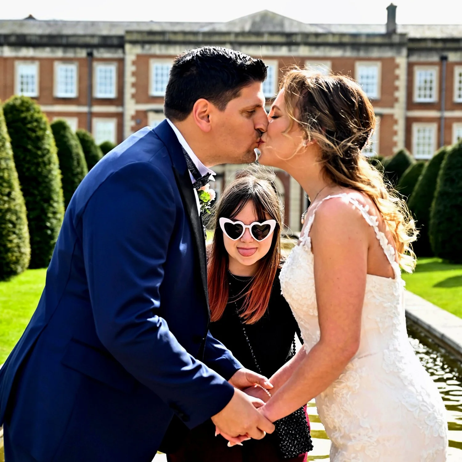 Candid wedding moment — bride and groom share a kiss while their neurodivergent daughter hilariously photobombs in heart shaped sunglasses — documentary wedding photography by Tasha Boorman, Nottinghamshire