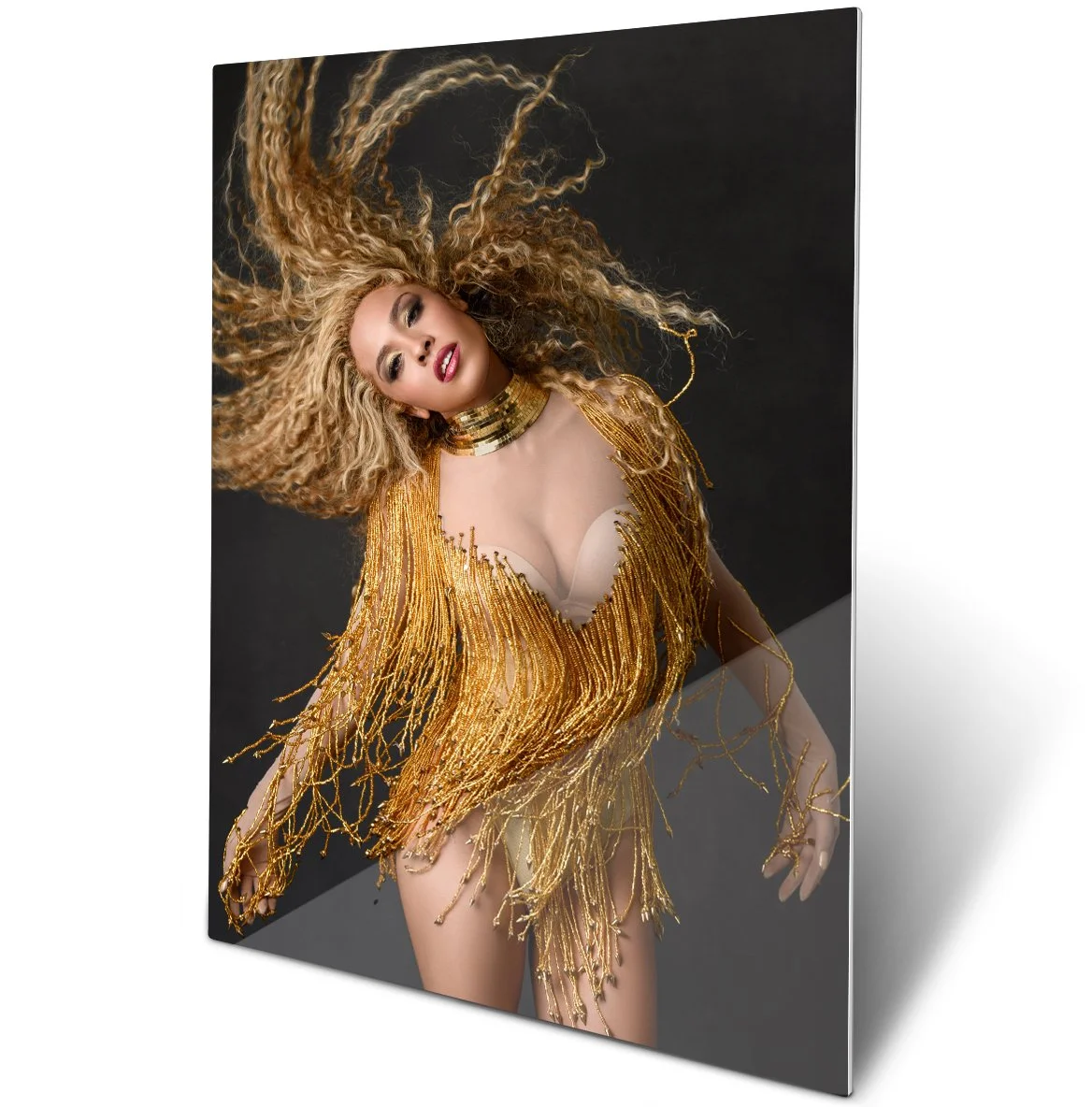 A woman with curly blonde hair posing in a gold fringed dress with a deep neckline and a wide metallic choker on black background.