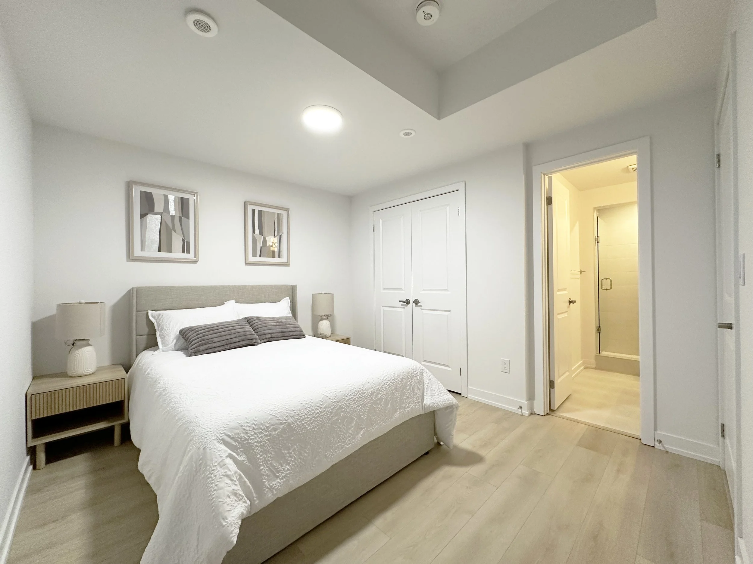 A minimalistic bedroom with a bed covered in white quilt, two matching beige nightstands with lamps, abstract art pieces on the wall, a closed closet door, and an open door leading to a bathroom with a shower.