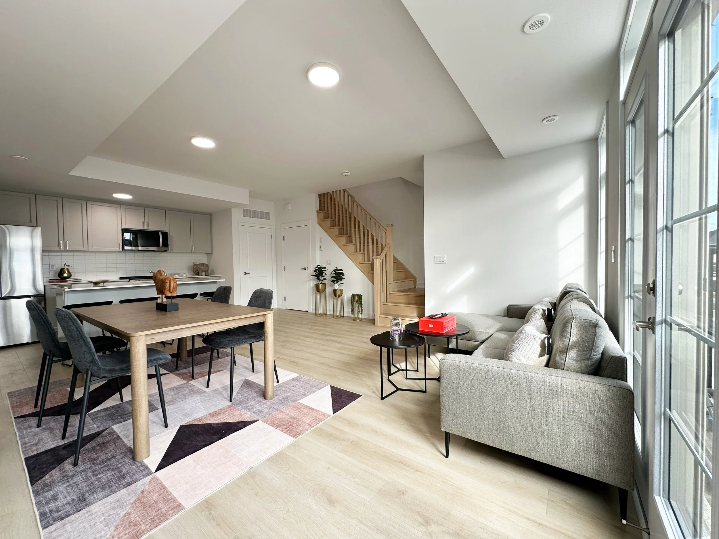 Open-concept living room and kitchen with large windows, staircase, gray sofa, dining table with chairs, and modern decor.