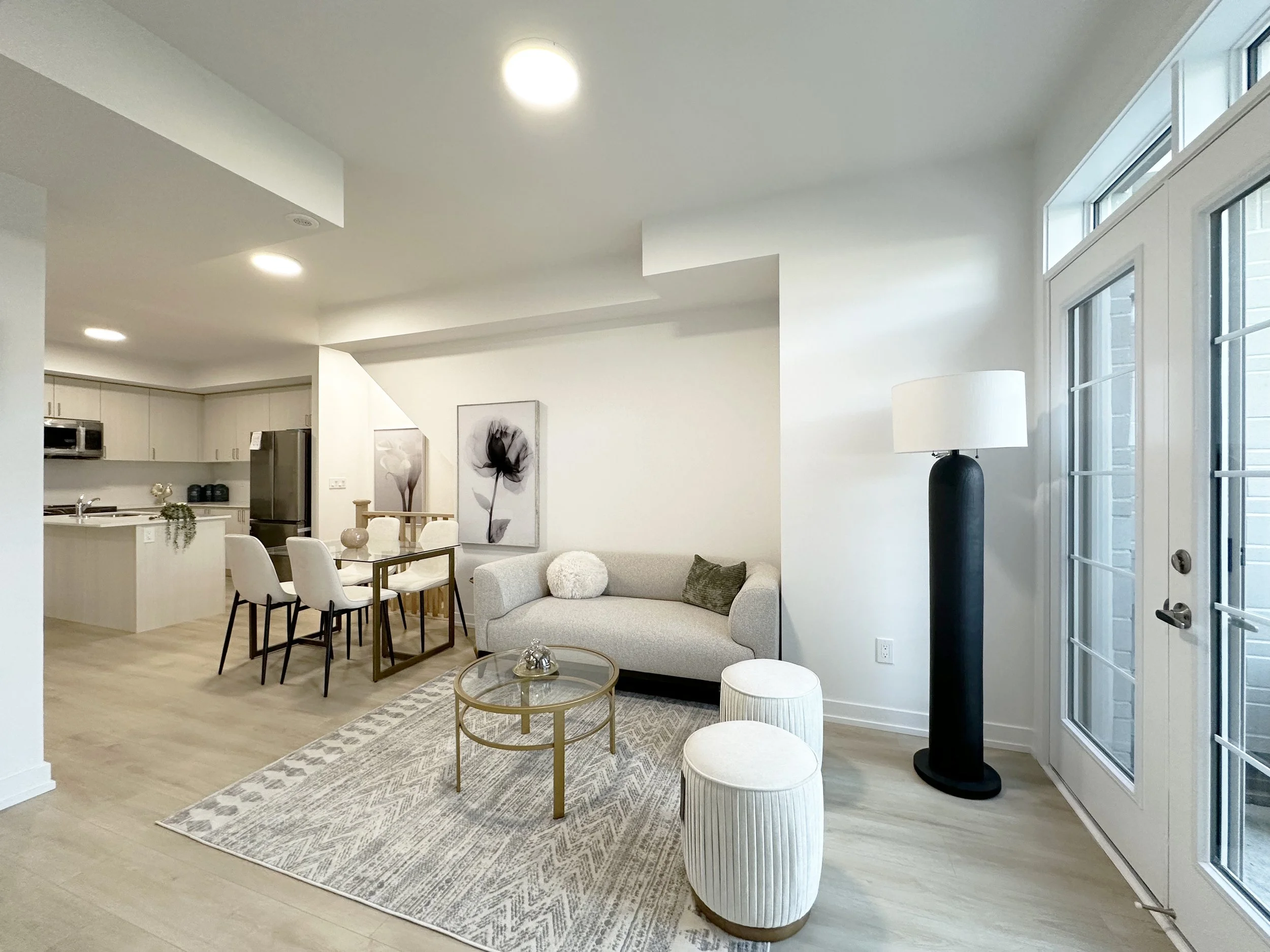 Living room with white walls, a gray sofa with pillows, a glass coffee table, a black floor lamp near a large window, and a dining area with white chairs and artwork on the wall, open to a kitchen.