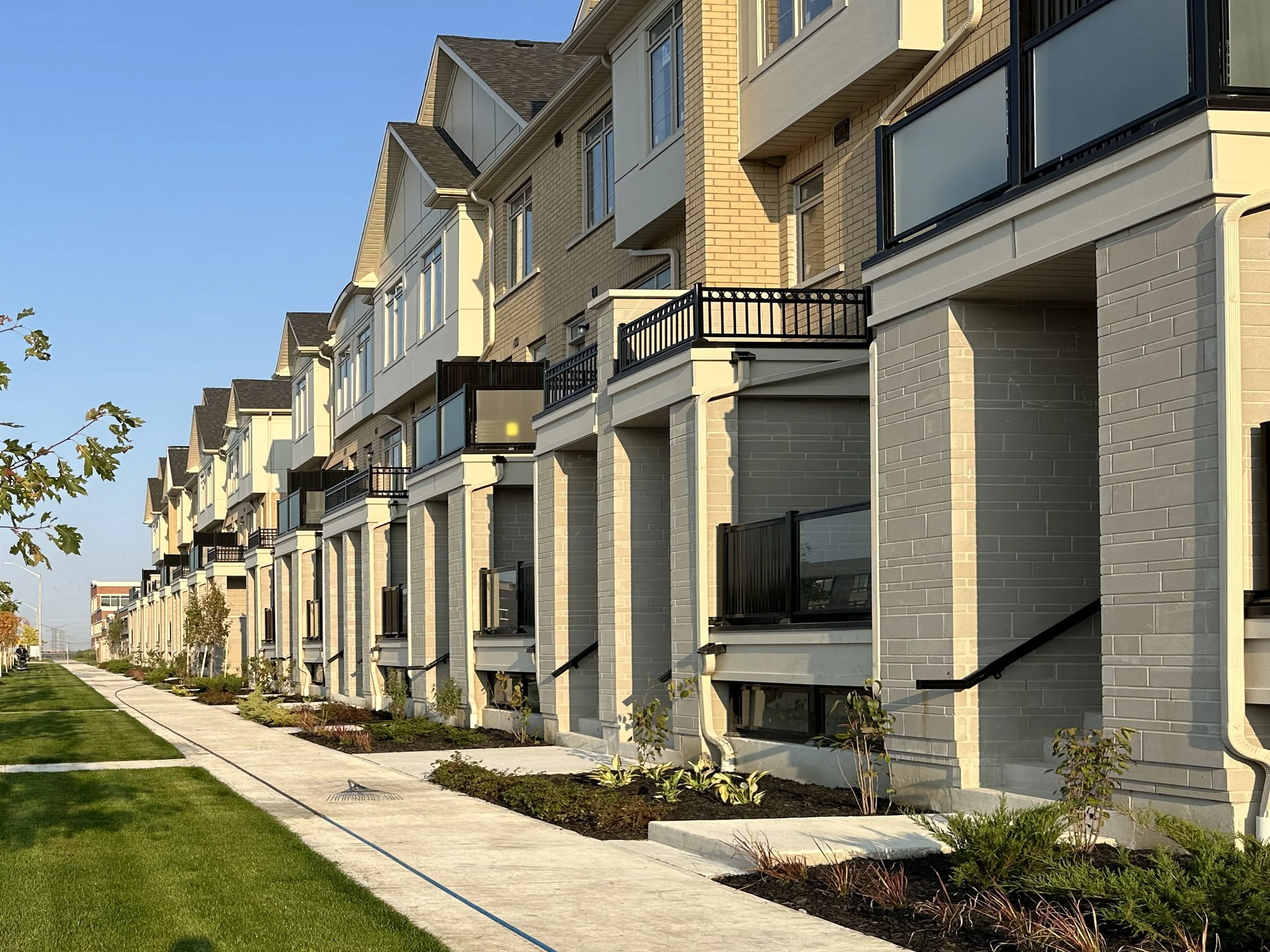 A modern apartment building with multiple floors, balconies with black railings, large windows, and a variety of siding and brick exterior finishes. The landscaping includes a well-maintained lawn, young plants, and a sidewalk along the building, und