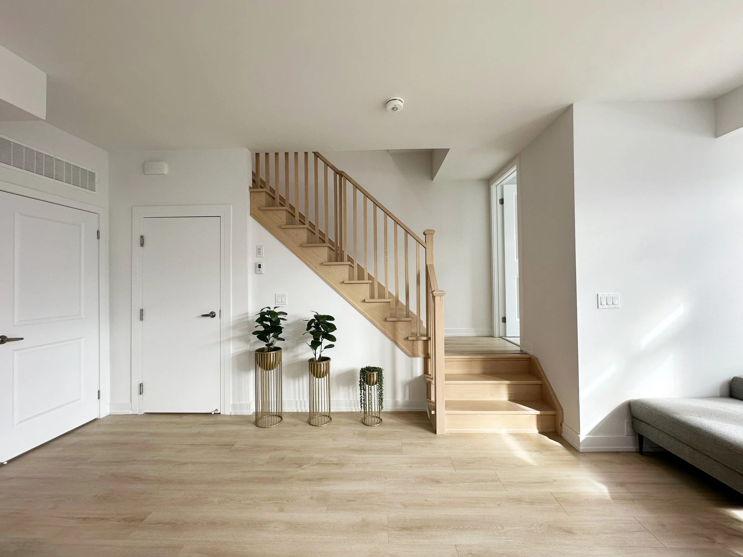 A modern living room with white walls, light wooden stairs, three gold-colored plant stands with green plants, a gray sofa, and a large window on the right.
