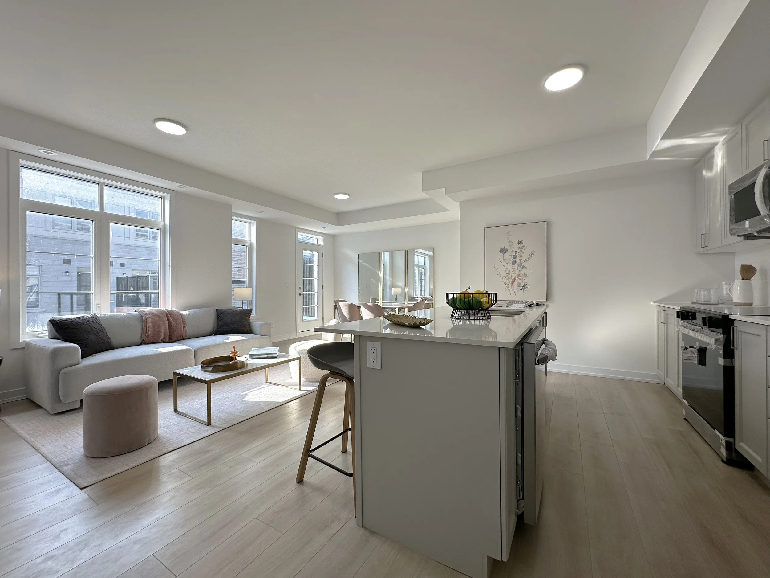 Open-concept living room and kitchen with large windows, a white sofa, round stool, kitchen island with fruit bowl, and modern appliances.