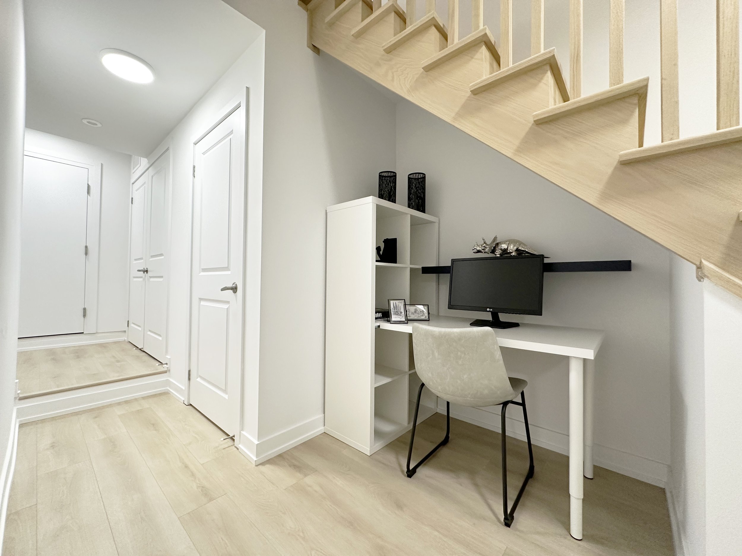 A modern, minimalist home workspace with a white desk, a beige upholstered chair, a computer monitor, and decorative items including framed pictures and black candle holders underneath a wooden staircase.