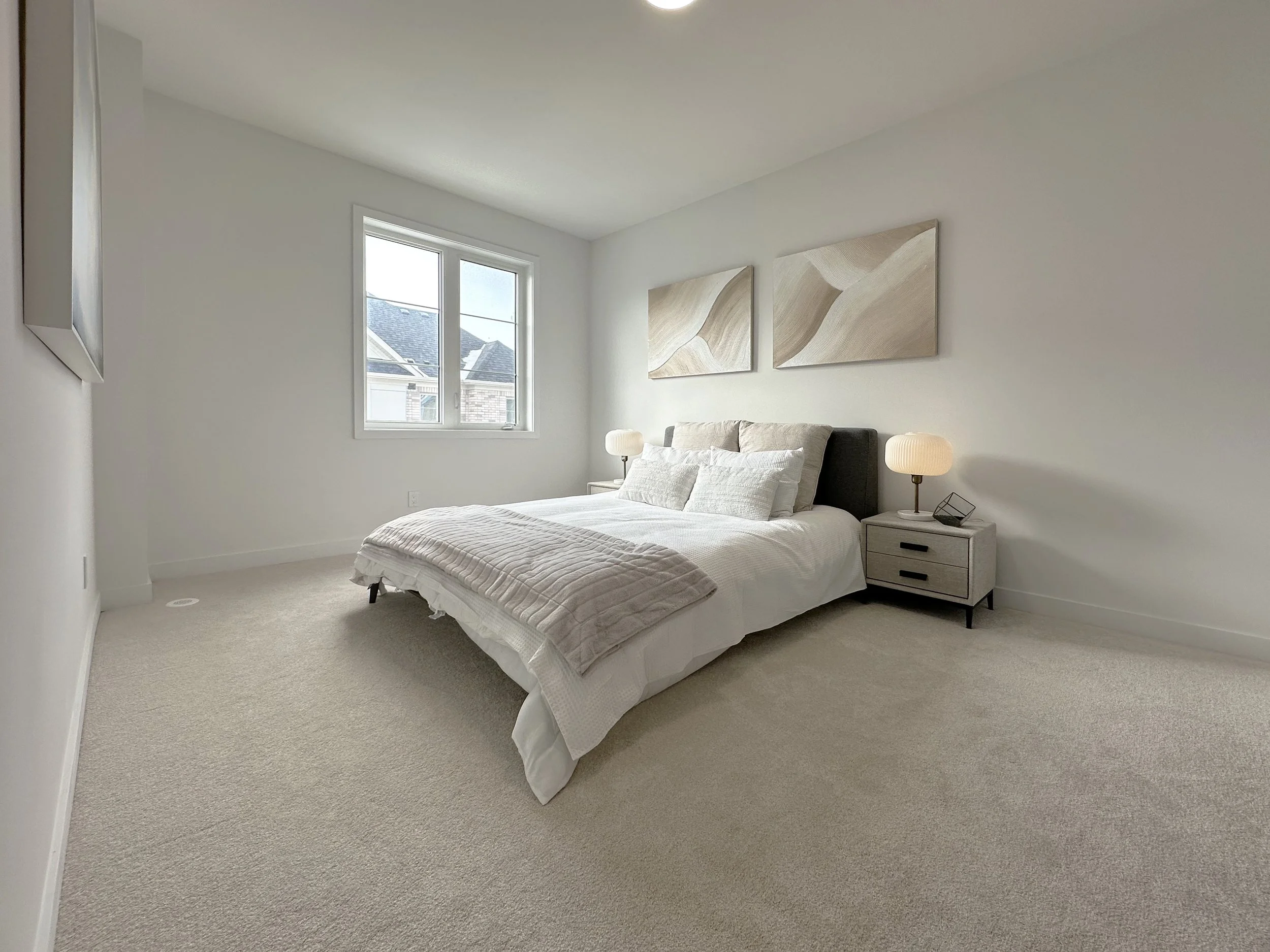 A minimalistic bedroom with a bed, nightstands, lamps, artwork, and a window showing rooftops outside.