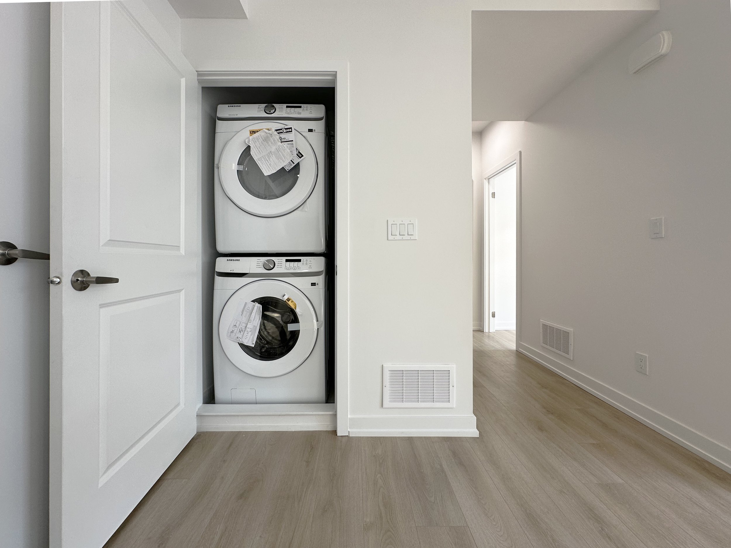 A small laundry area with a stacked washer and dryer, enclosed within a closet with a partially open door. The surrounding walls are painted white, and the floor features light-colored wood or laminate flooring. There is a vent on the wall near the f