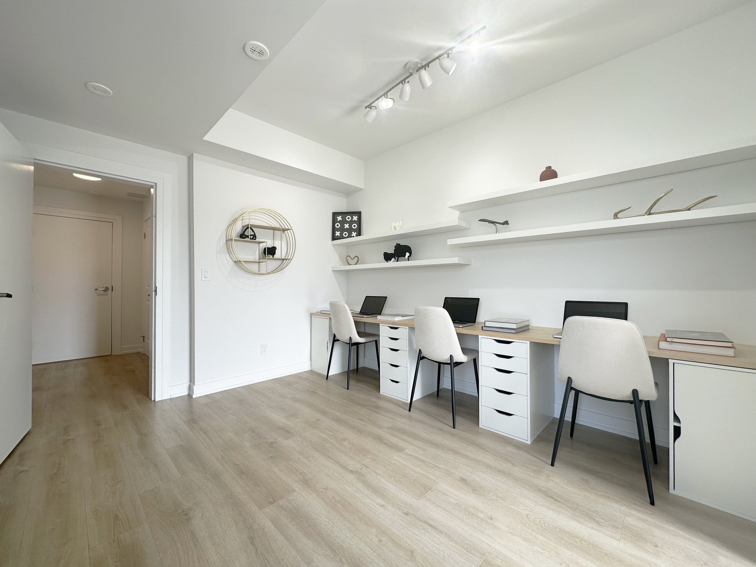 A bright, modern home office with beige chairs at a long white desk, white walls, floating shelves decorated with black and gold accents, and wood flooring.