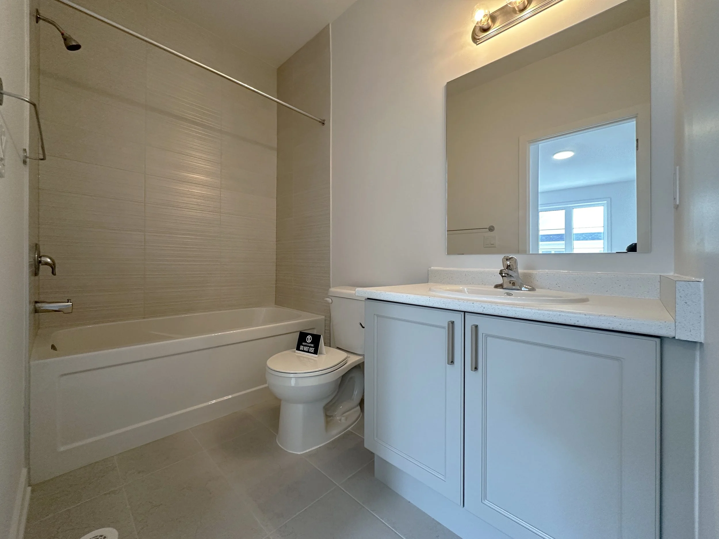 Bathroom with a bathtub, shower head, toilet with a sign that says 'Do Not Use', white vanity with a sink, mirror, and window, beige floor tiles, and bright lighting