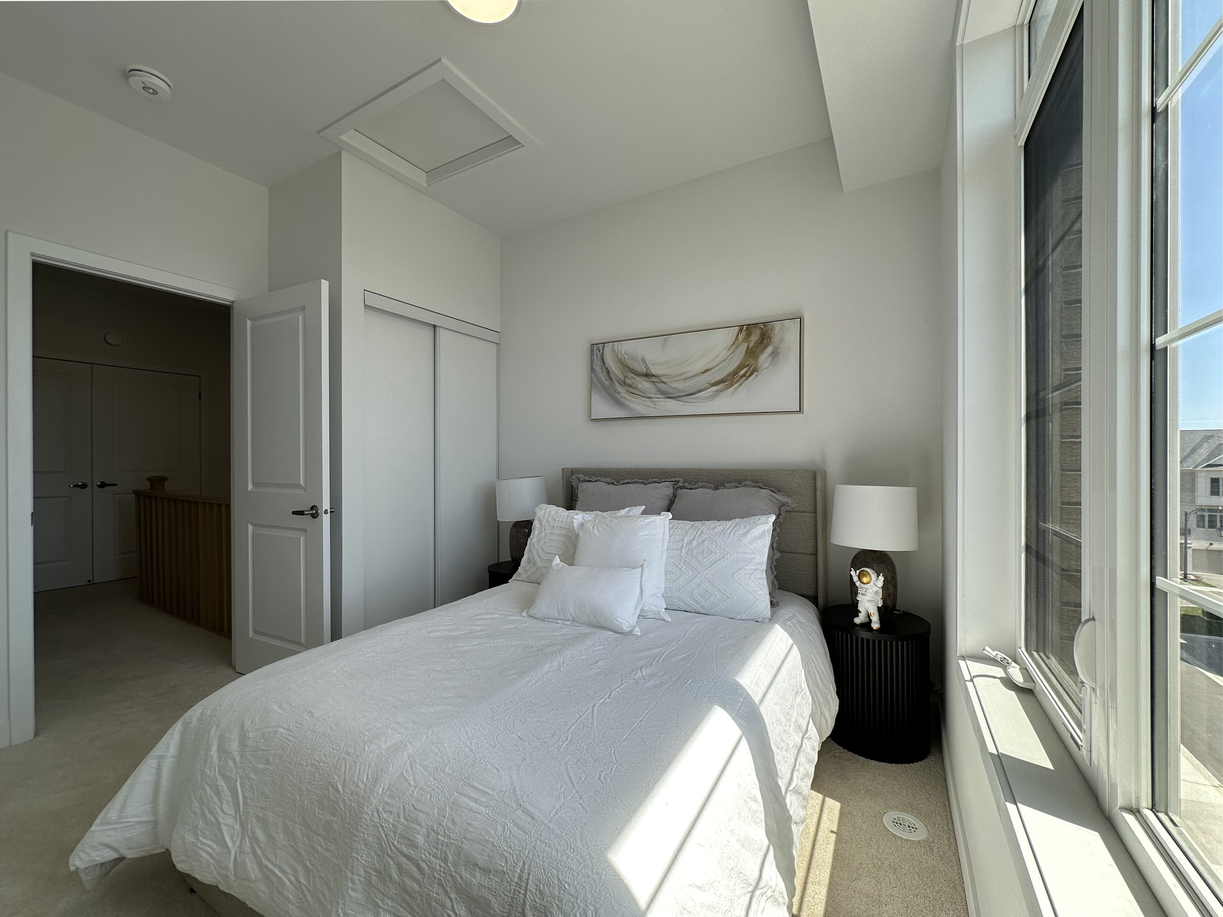 A bedroom with a white bed, pillows, nightstand with a lamp and astronaut figurine, large window with sunlight, artwork on the wall, and an open door leading to a closet.