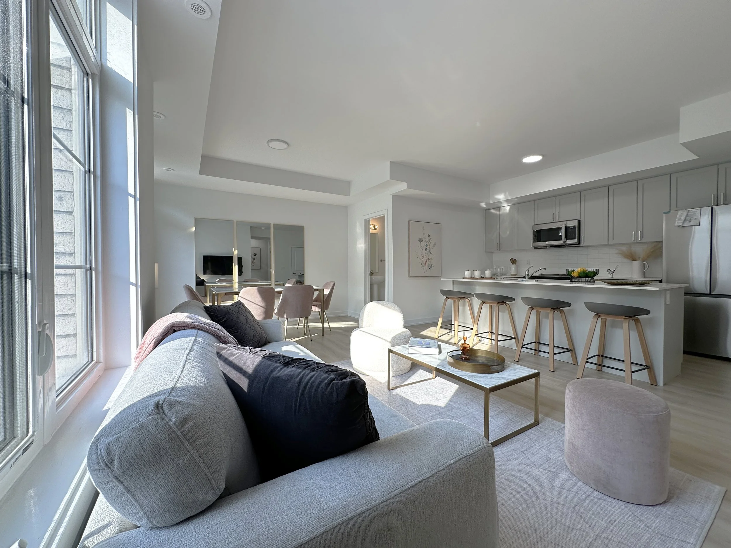 Living room and kitchen with large windows, beige sofa with dark cushions, round side table, kitchen with gray cabinets, white backsplash, stainless steel microwave, and refrigerator, bar stools, and a dining area in the background.