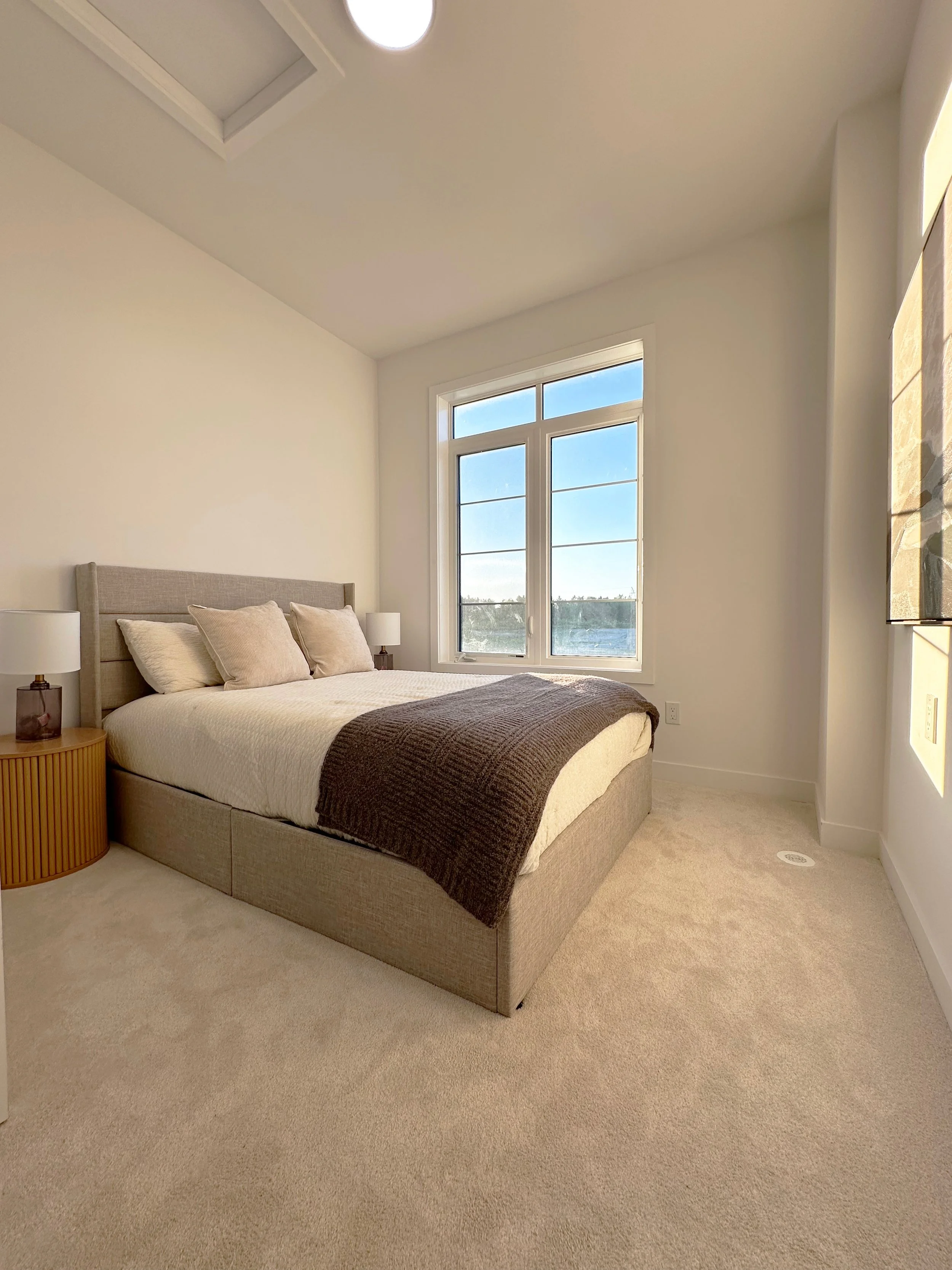 A bedroom with a bed, beige upholstered headboard, white bedding, brown blanket, beige pillows, two white table lamps on wooden nightstands, large window with a lake view, beige carpet, and a ceiling light.