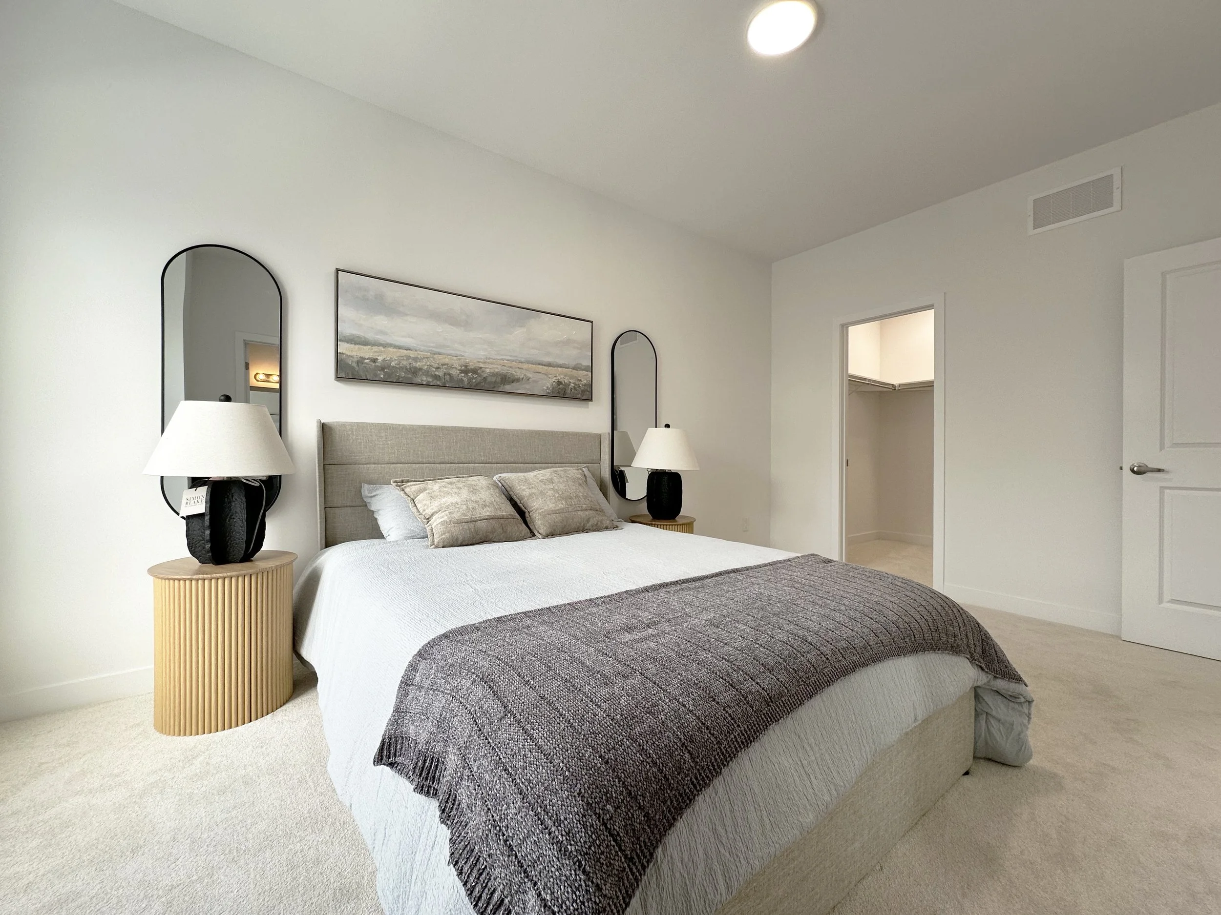 A bedroom with a bed, two lamps on bedside tables, and a landscape picture on the wall. The room has white walls and a beige carpet.