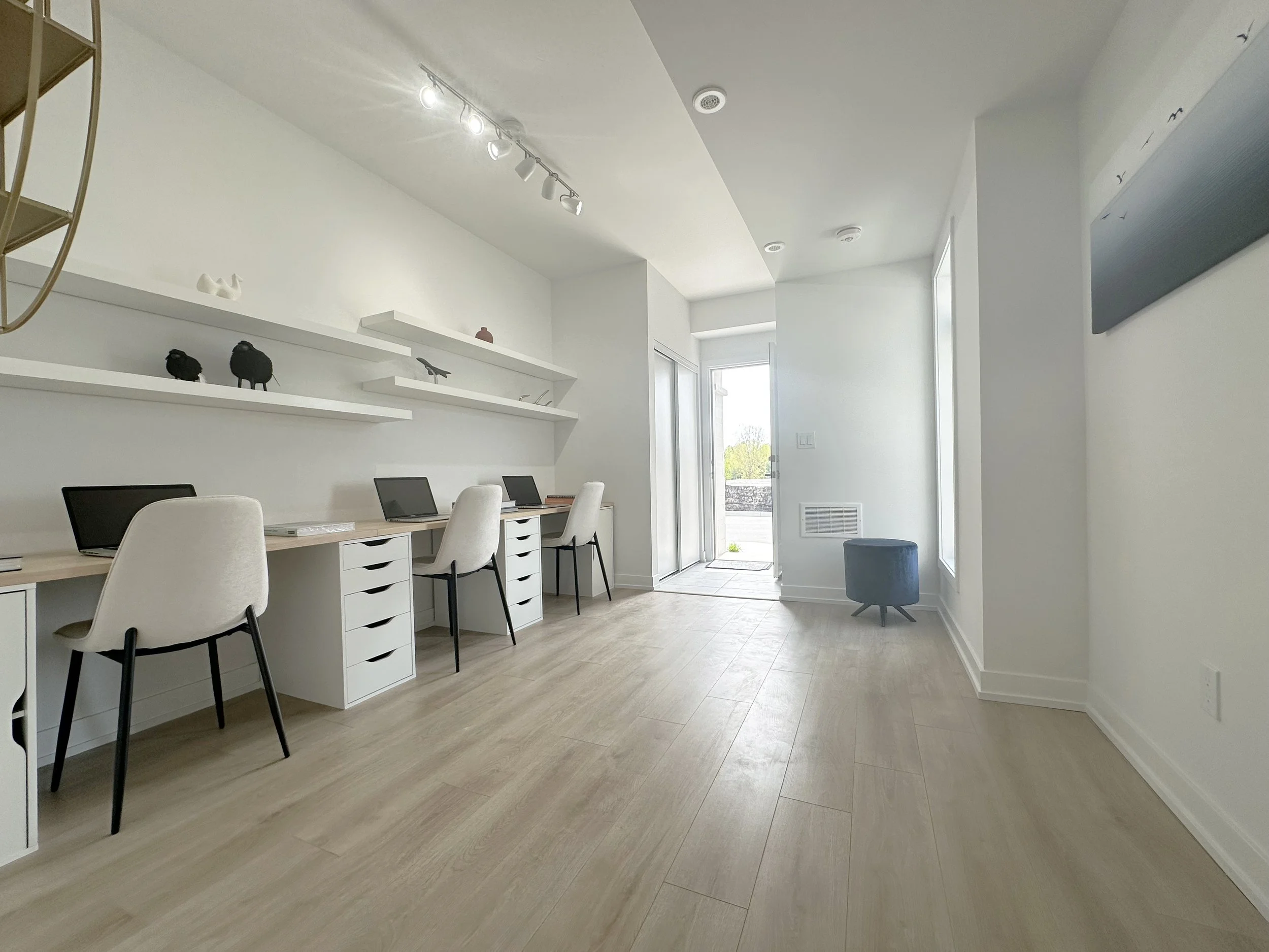 Brightly lit modern interior space with a long white desk, laptops, white chairs, white floating shelves with decorative objects, large windows, and light wood flooring.