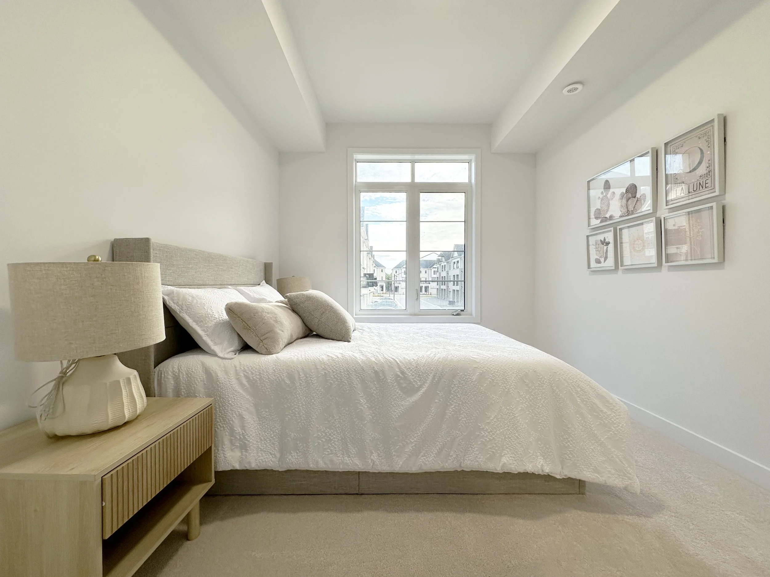 A bedroom with a bed, bedside table with a lamp, large window showing buildings outside, and framed art on the wall.