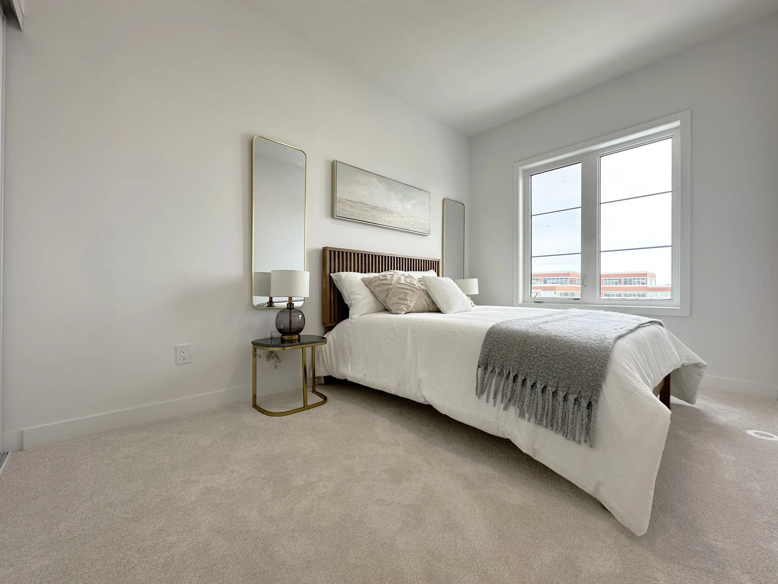A minimalist bedroom with a large window, beige carpet, a bed with white linens, decorative pillows, a wooden headboard, a small side table with a lamp, and two tall mirrors on the wall.