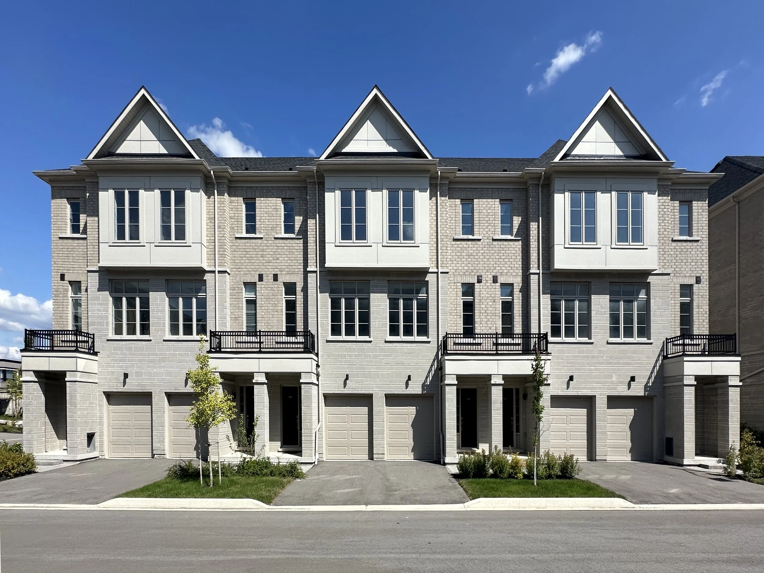 Front of a modern multi-story residential building with multiple garage doors, small trees, and a sidewalk, under a clear blue sky.