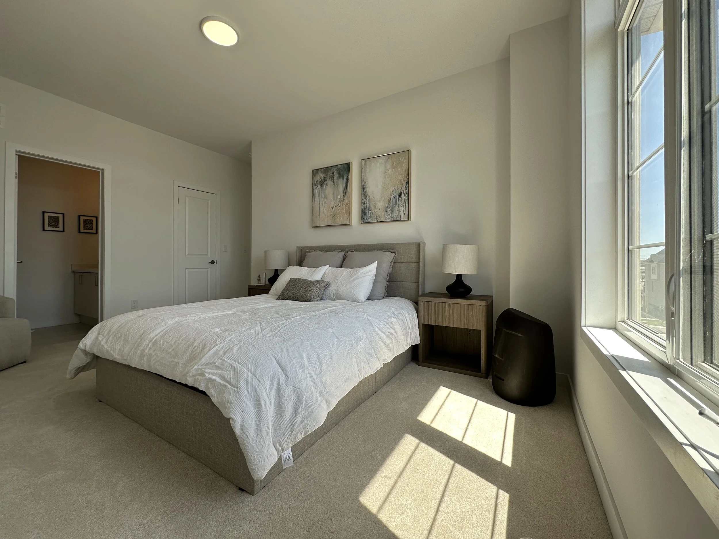 A bedroom with a large bed, nightstands with lamps, and abstract wall art, illuminated by sunlight coming through large windows.