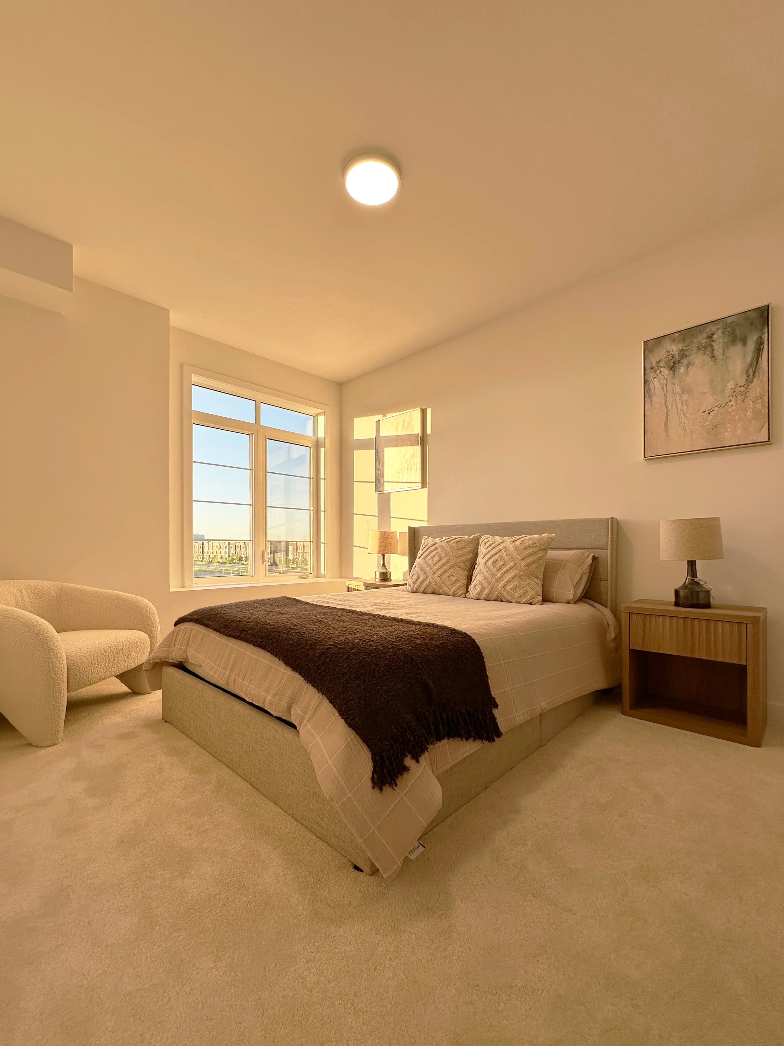 A bedroom with a beige bed, pillows, side table, lamp, armchair, large windows, and wall art.