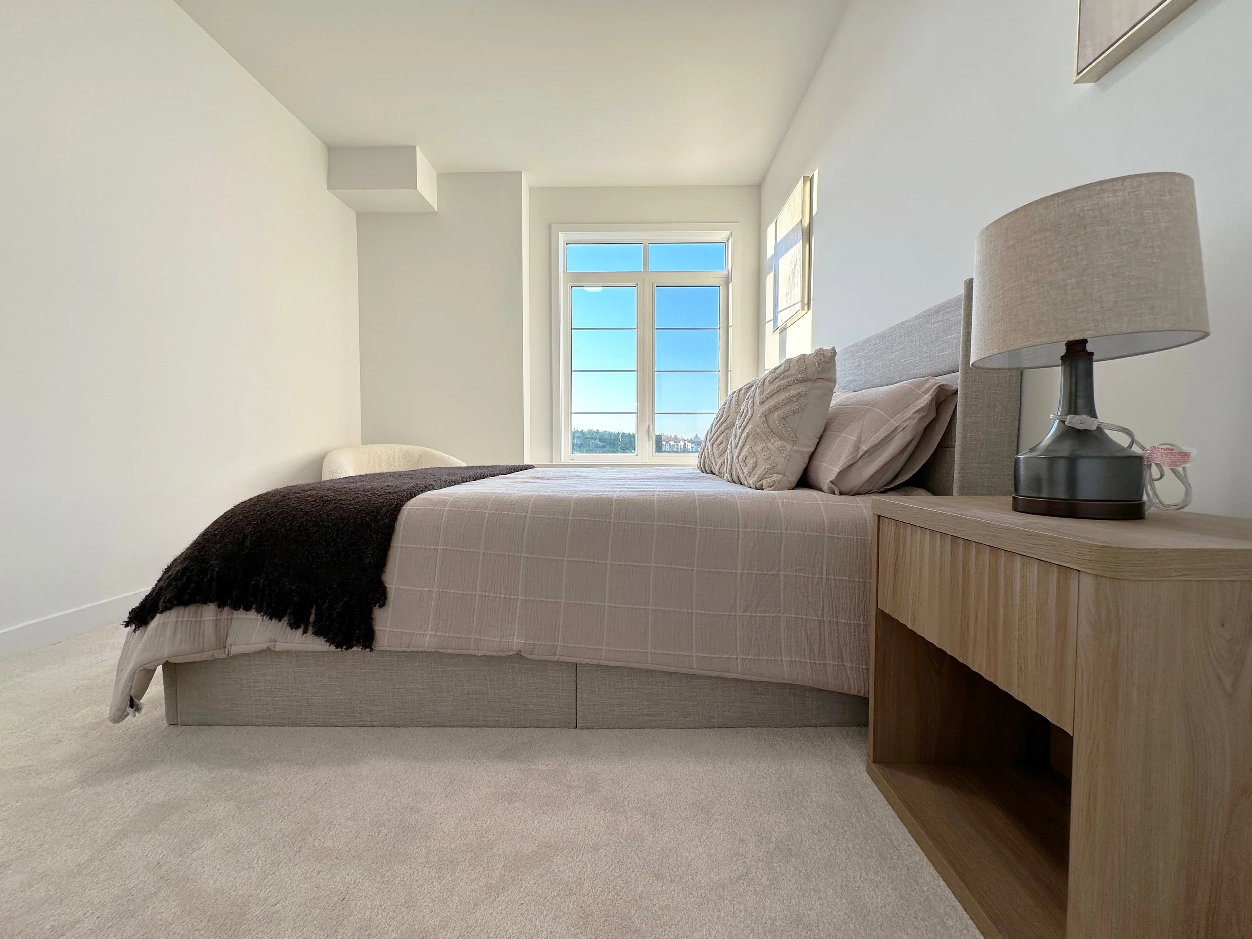 A bedroom with a bed that has beige linens, multiple pillows, and a dark brown throw blanket. There is a wooden nightstand with a lamp and some items on it. Large windows with a clear blue sky visible outside, and beige carpet flooring.