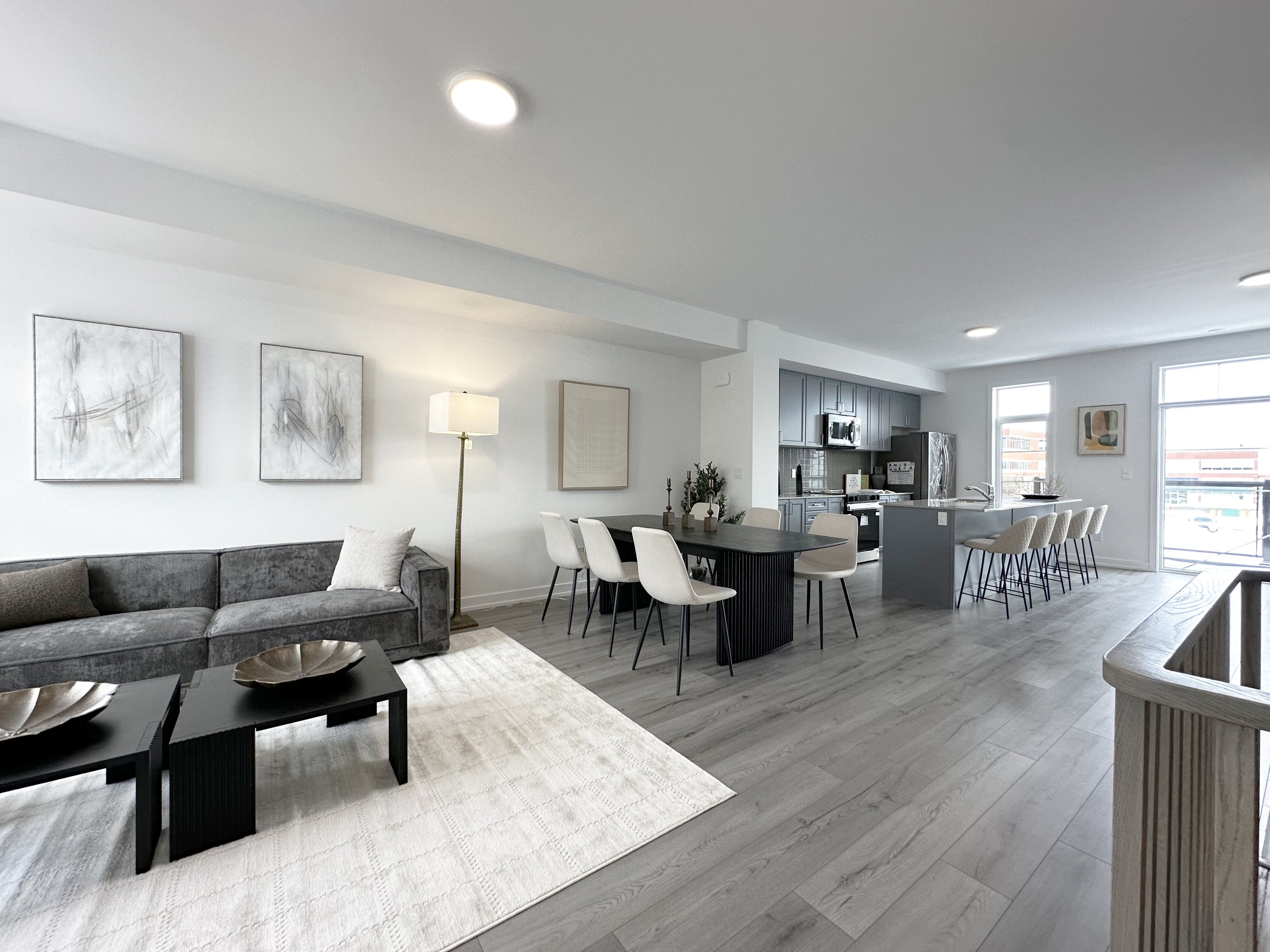 Modern open concept living room and kitchen with gray cabinetry, white walls, and large windows, furnished with a gray sofa, white chairs, and black coffee tables.