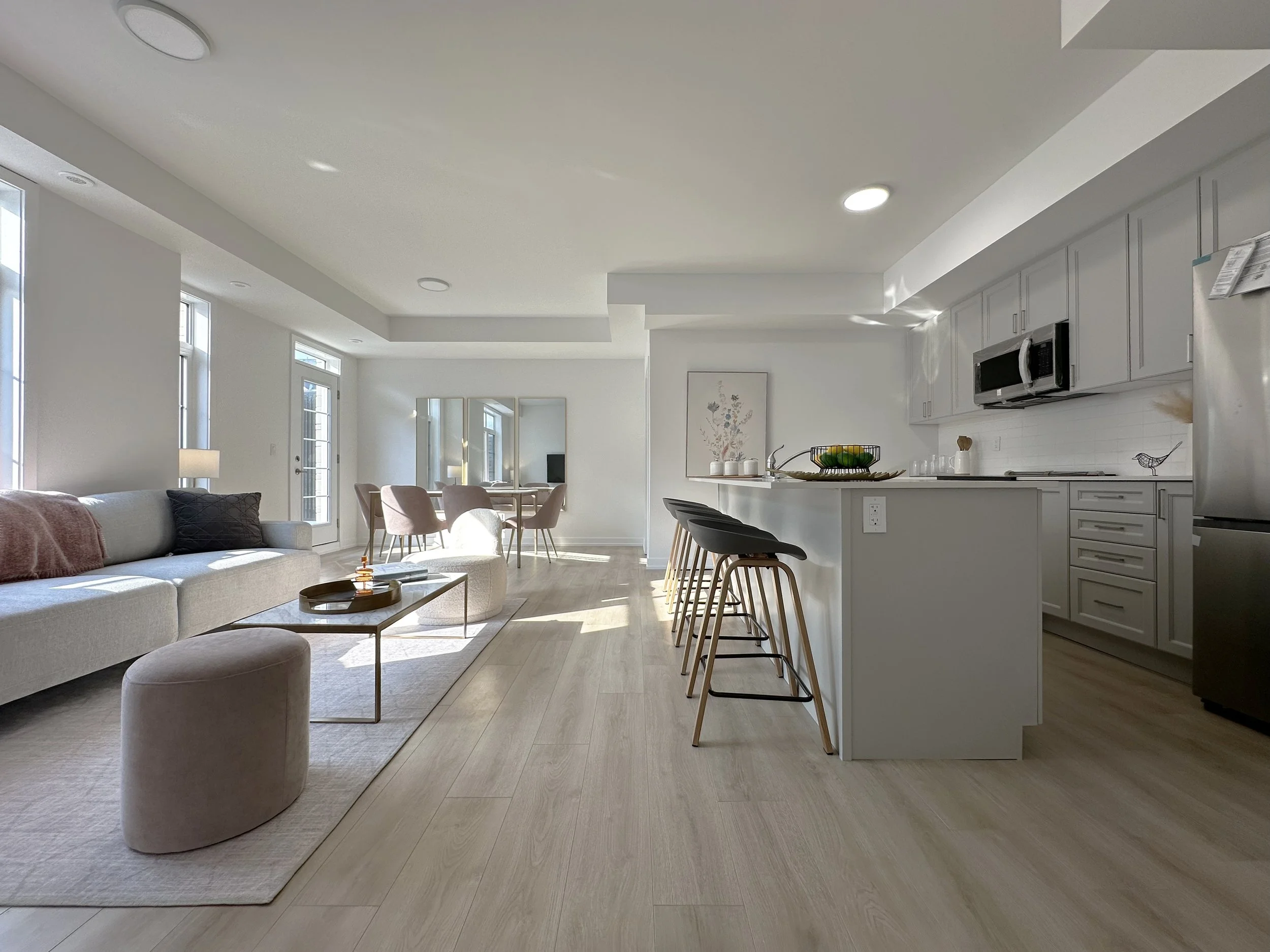 Open-concept living room and kitchen with light wood flooring, white walls, large windows, a gray sectional sofa, a round ottoman, a rectangular coffee table, a dining table with chairs, and kitchen cabinetry with appliances.