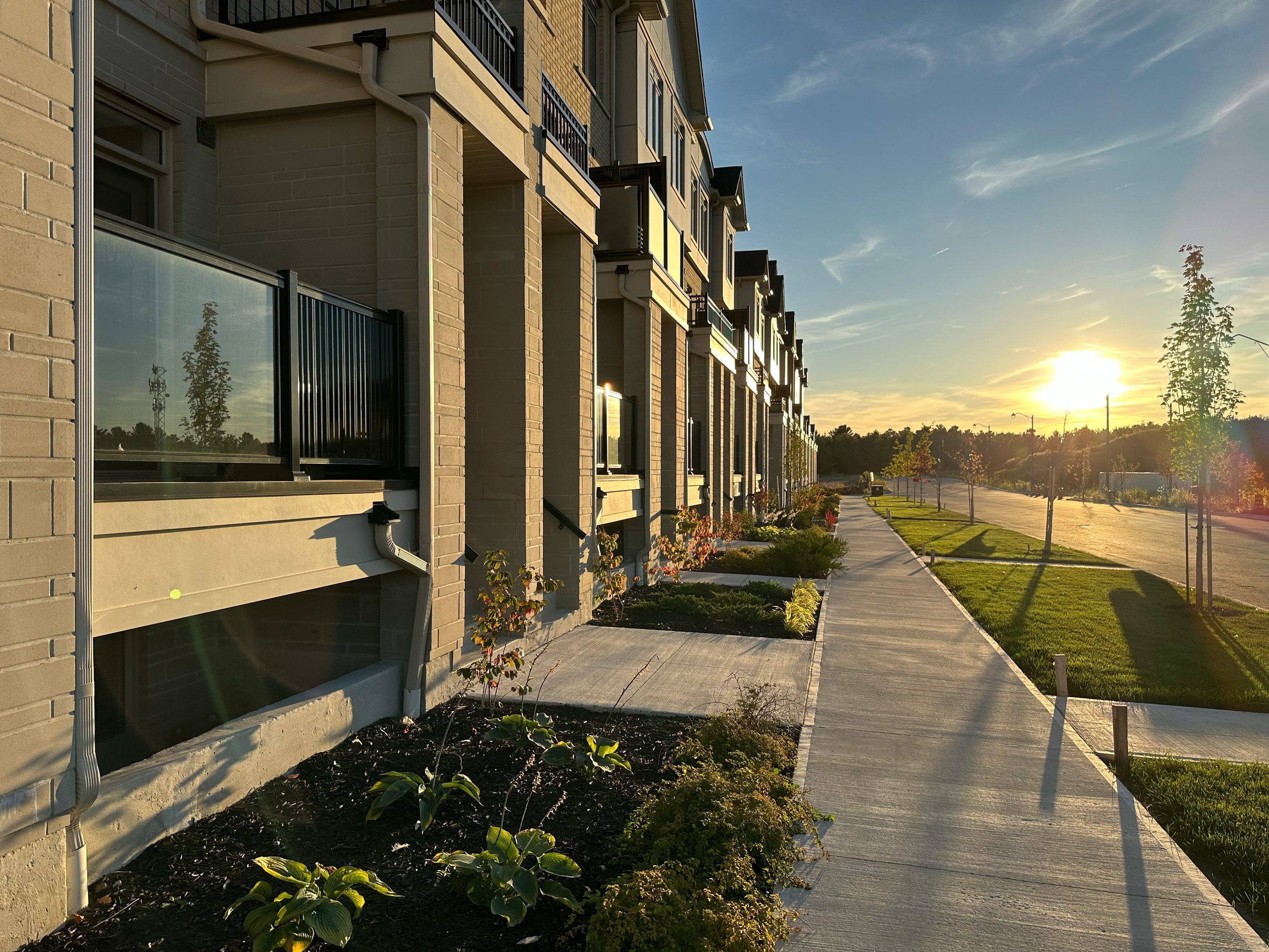 Sidewalk in front of modern apartment building with small plants and trees, sunny weather, blue sky with some clouds.