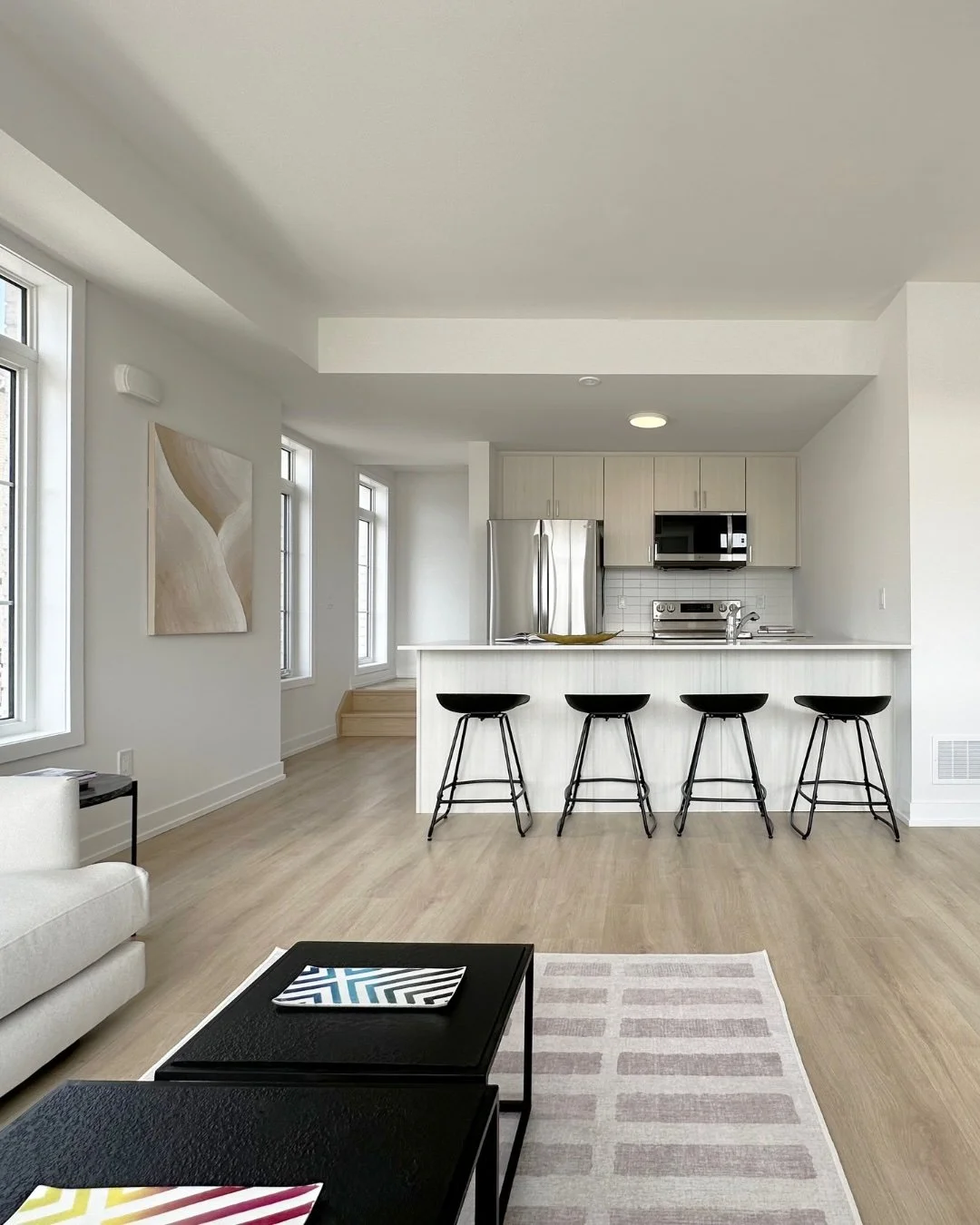 Modern kitchen with light wood cabinets, stainless steel refrigerator, microwave, and stove. Kitchen island with four black bar stools. Light-colored hardwood floors, white walls, and large windows with natural light. Living area visible with a white sofa, black coffee tables, and a patterned rug.