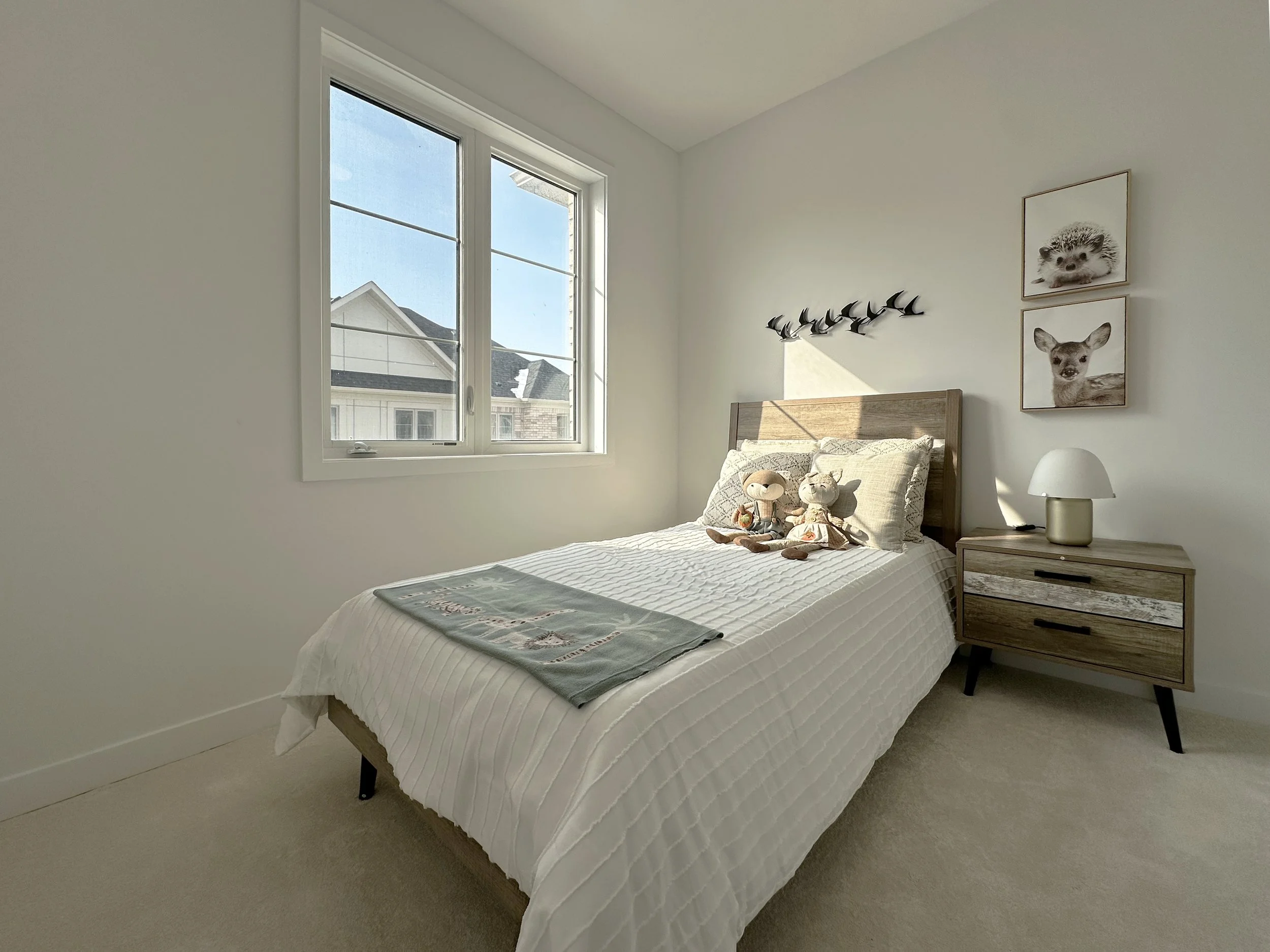 A bedroom with a large window, a bed with a white quilted bedding, and a wooden headboard. On the bed are two teddy bears, one larger and one smaller. To the right of the bed is a wooden nightstand with a white lamp. The wall above the bed has two fr