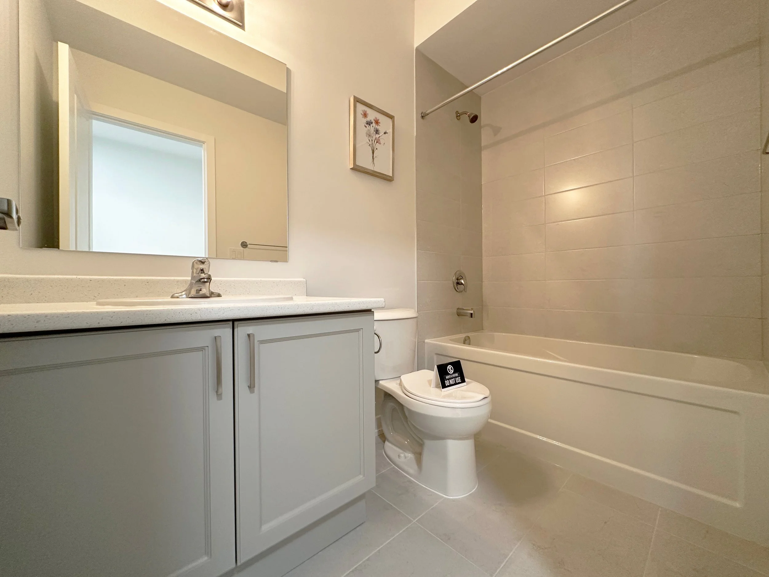 A bathroom with a white vanity sink, large mirror, framed flower picture on the wall, bathtub with a shower curtain rod, and a toilet with a "Do Not Use" sign on the lid.