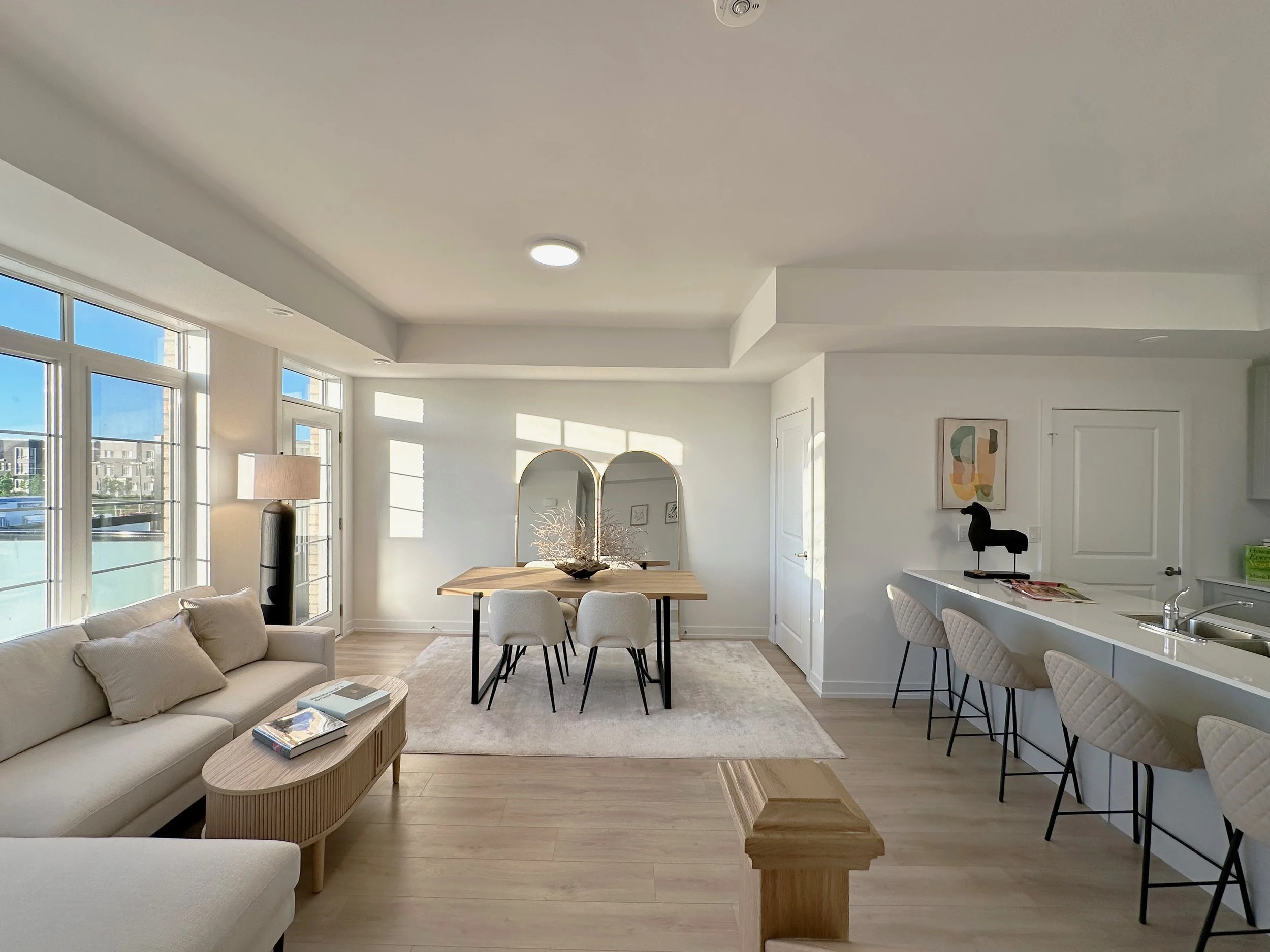 Modern open-plan living and dining area with large windows, white walls, a beige sofa with pillows, a wooden dining table with chairs, a light-colored rug, and decorative items including a vase with dried branches and framed artwork.