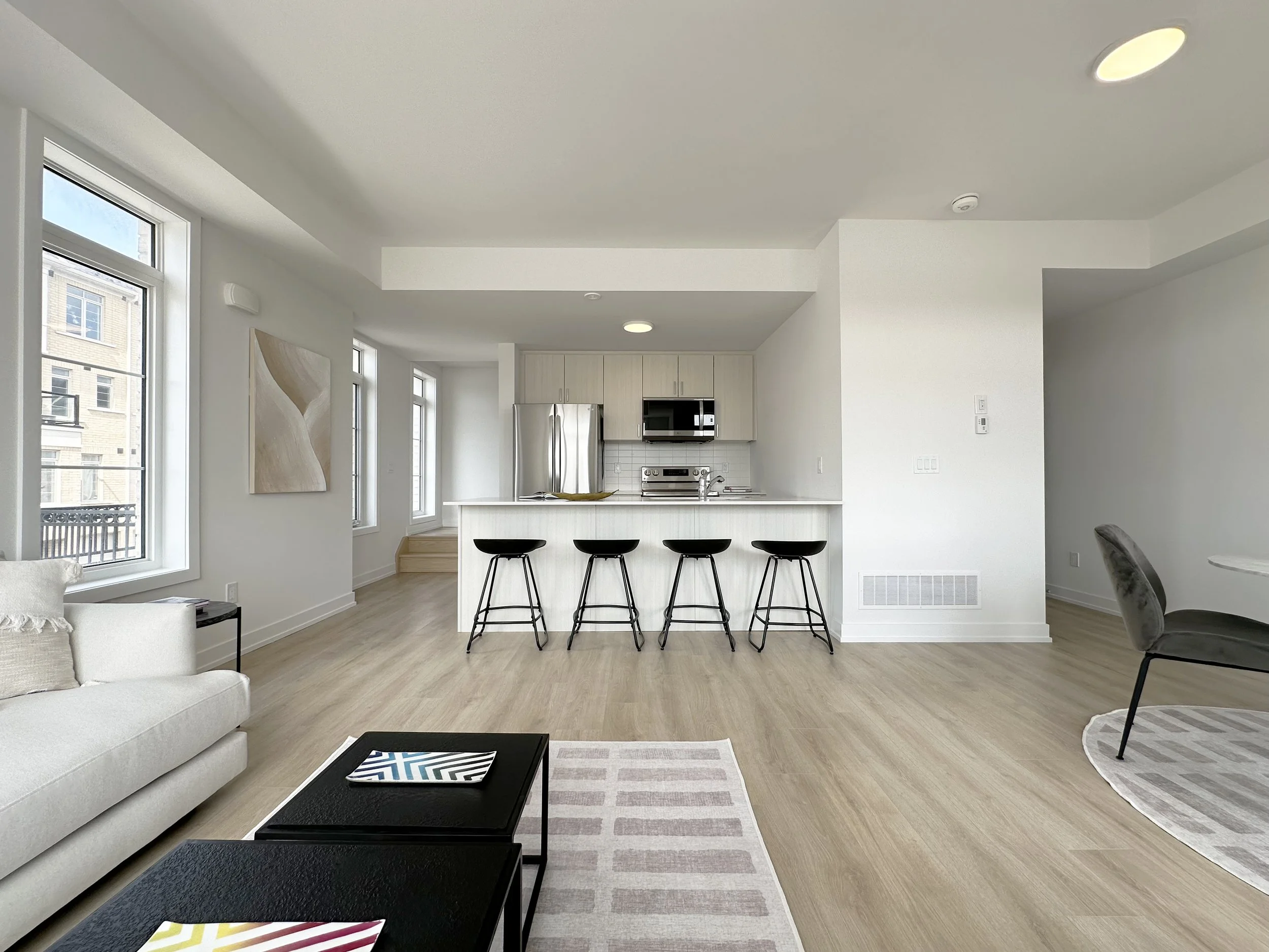 Open-concept living room and kitchen in a modern apartment with large windows, light-colored walls and flooring, a white sofa, black coffee table, and kitchen island with four black barstools.