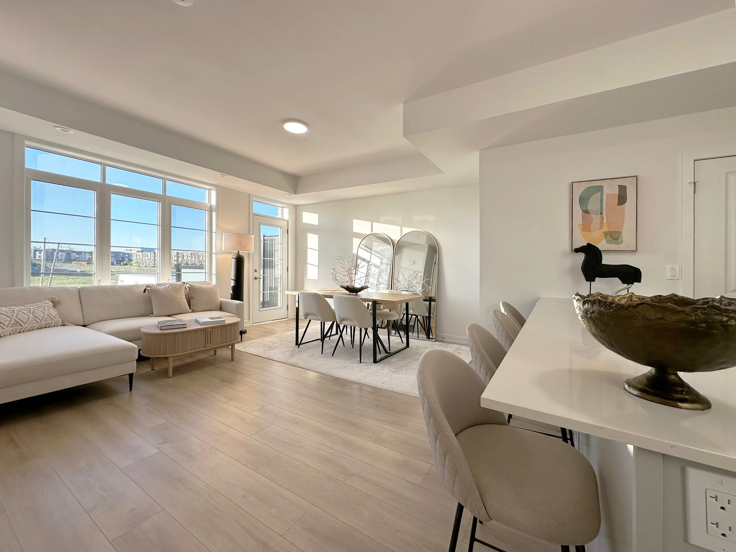 Spacious, modern living room with large windows, white walls, and light wood flooring. Contains a white sofa, a round coffee table, a lamp, a dining table with chairs, decorative art, and a sculpture on the table.