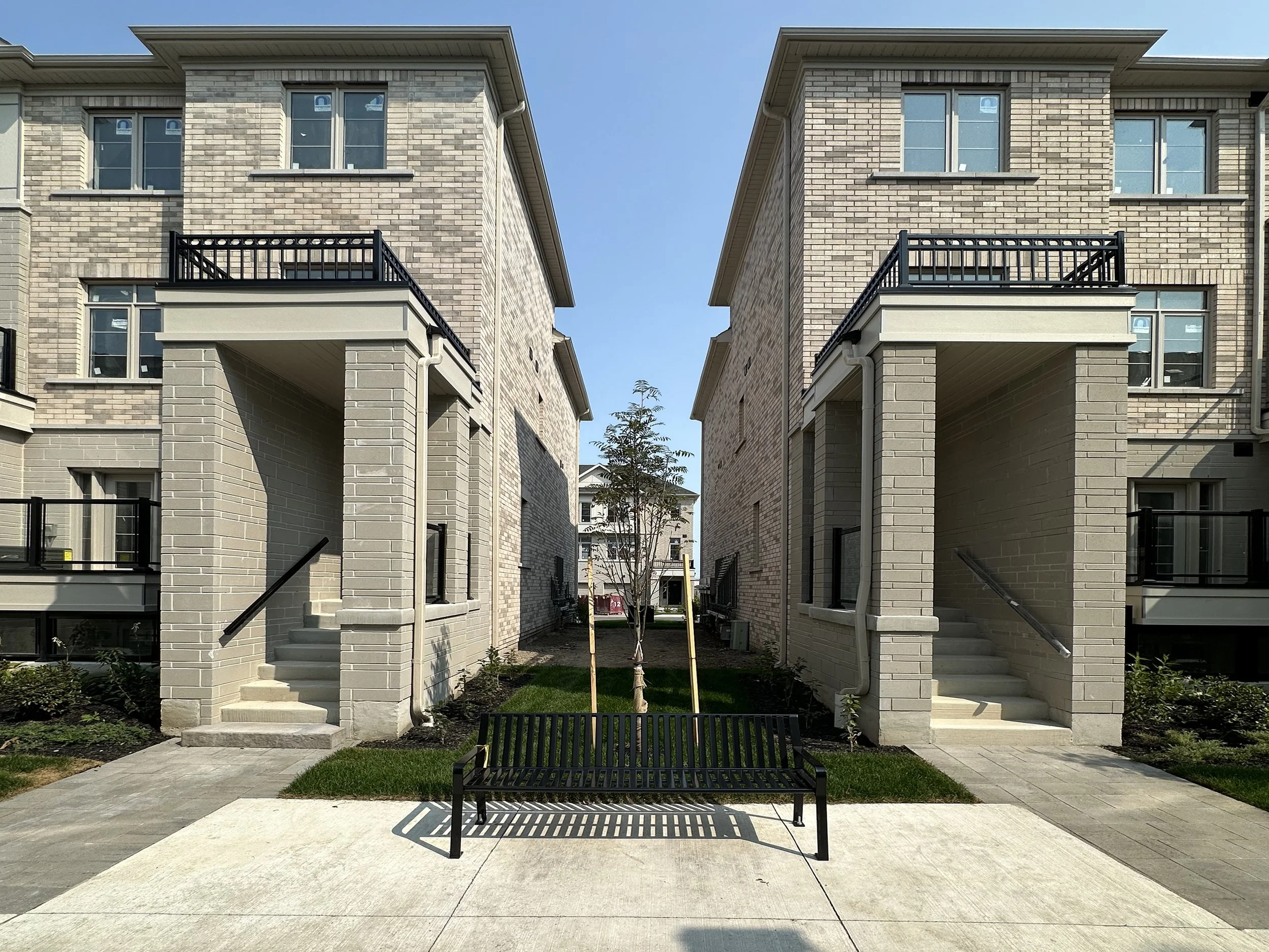 View of two modern apartment buildings with brick facades, featuring balconies with black railings. A pathway with steps leads to the buildings, and a young tree is planted in the courtyard area between them. The sky is clear and blue.