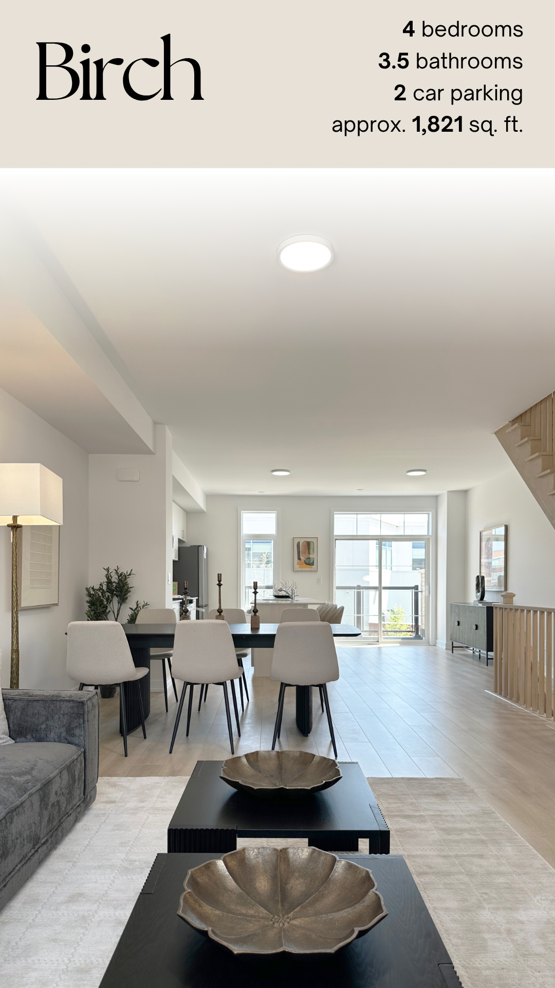 Open-concept living and dining area with white walls and light wood flooring, featuring a dark dining table with six white chairs, two black coffee tables with metallic bowls, a gray sofa, and large windows with natural light.