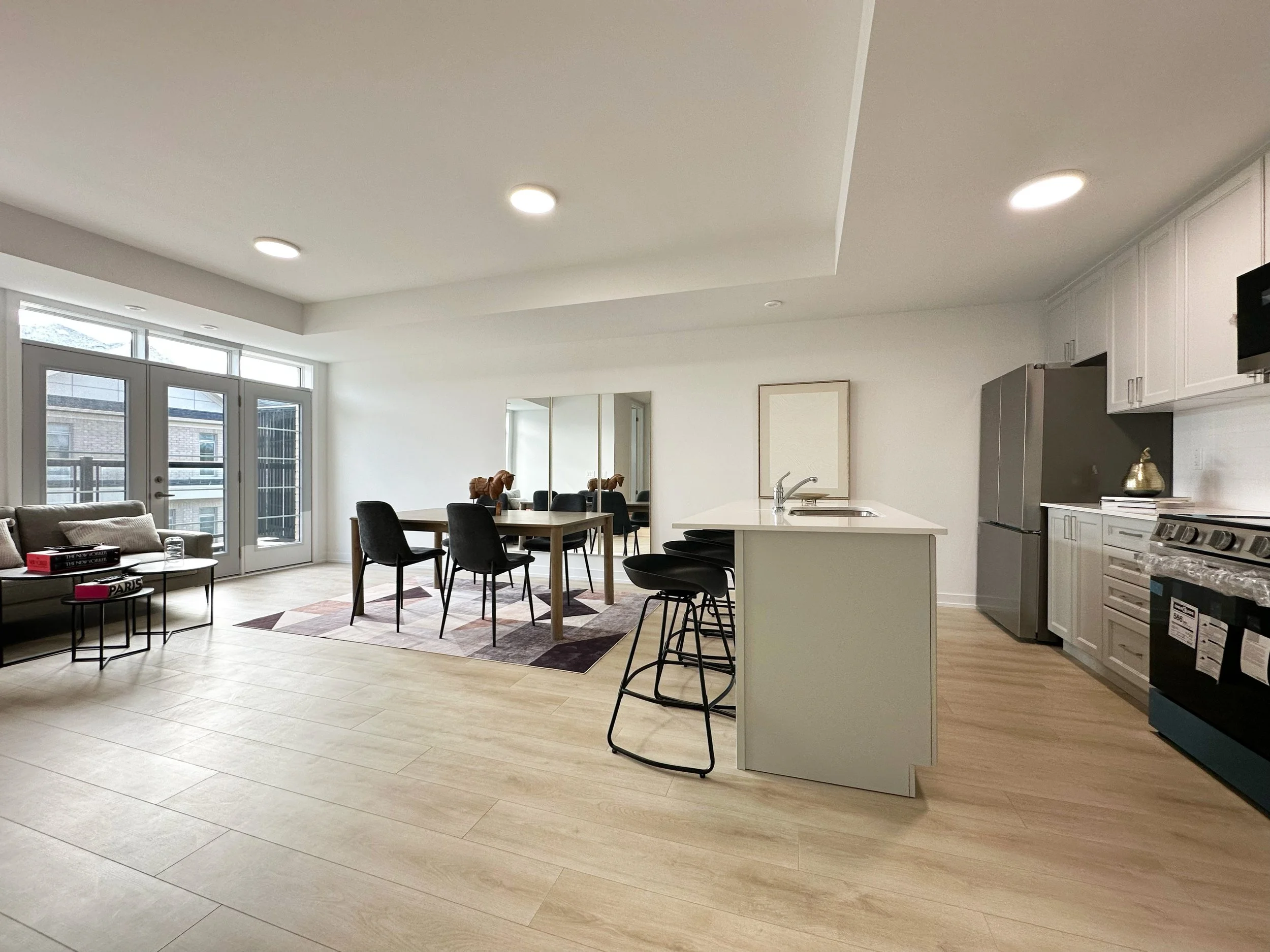 Open-concept living space with large windows, a dining area with a wooden table and black chairs, a white kitchen island, and white cabinetry; modern decor with framed artwork and a mirror, light wood flooring, and recessed lighting.