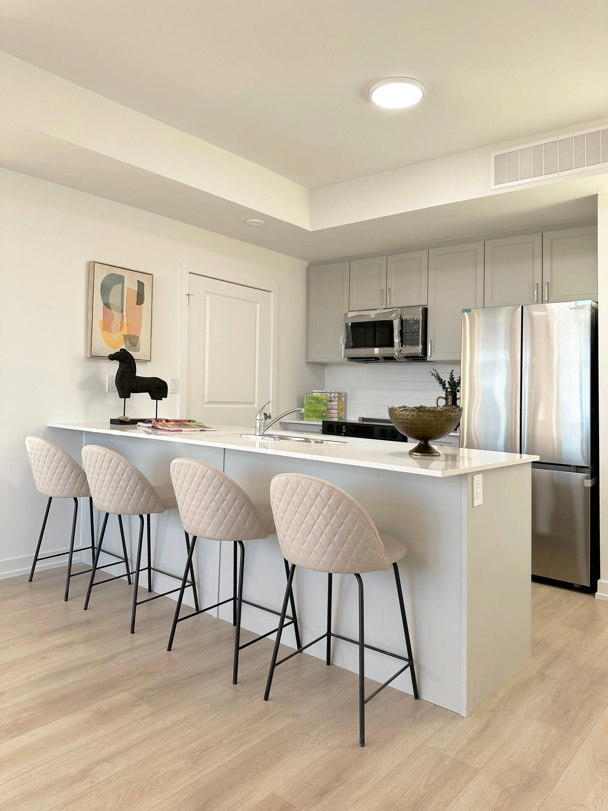 Kitchen with four beige quilted bar stools at a white island, gray cabinets, stainless steel refrigerator, built-in microwave, decorative bowl, and artwork on the wall.