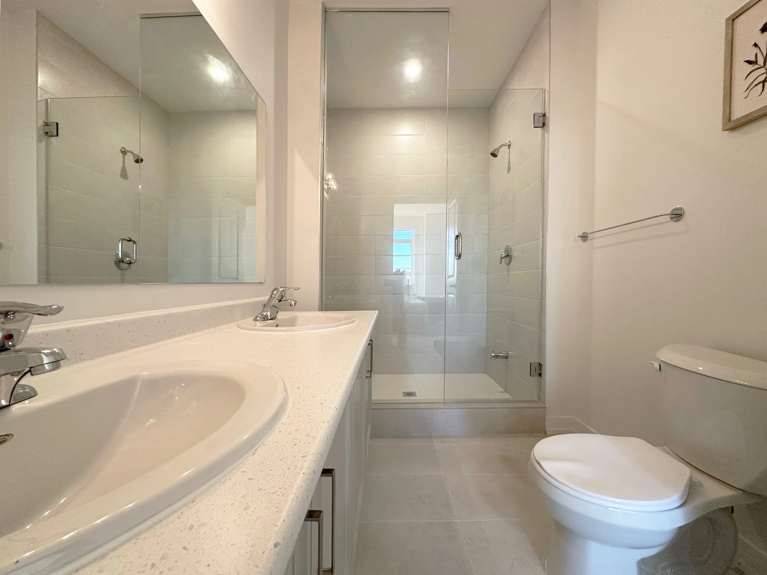 A modern bathroom with a white countertop, two sinks, a large mirror, a glass-enclosed shower, a white toilet, and wall art.