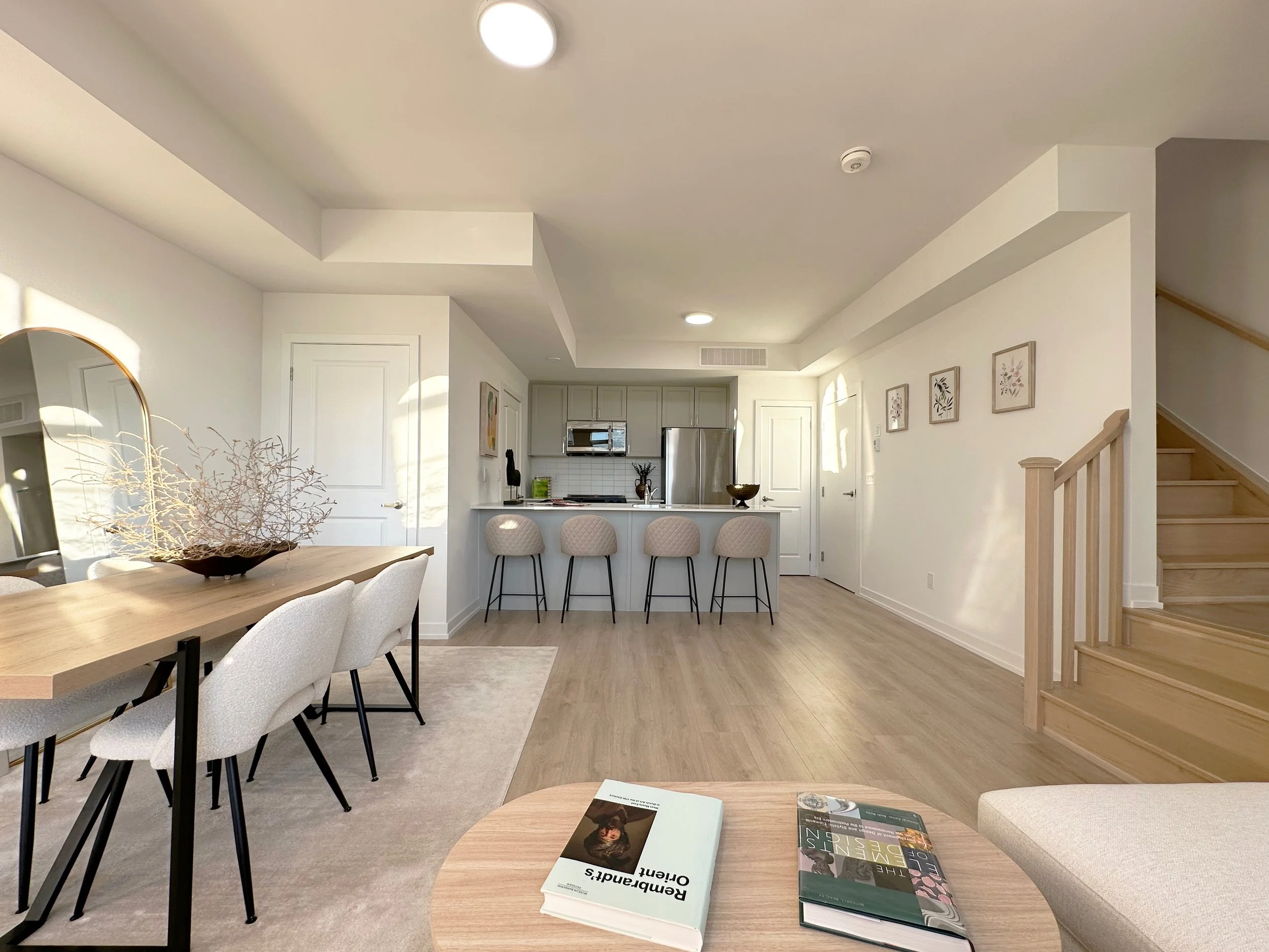 Interior view of a modern kitchen and dining area with white walls, light wood flooring, a dining table with white chairs, a kitchen counter with bar stools, and artwork on the walls. There are books on a round table in the foreground.
