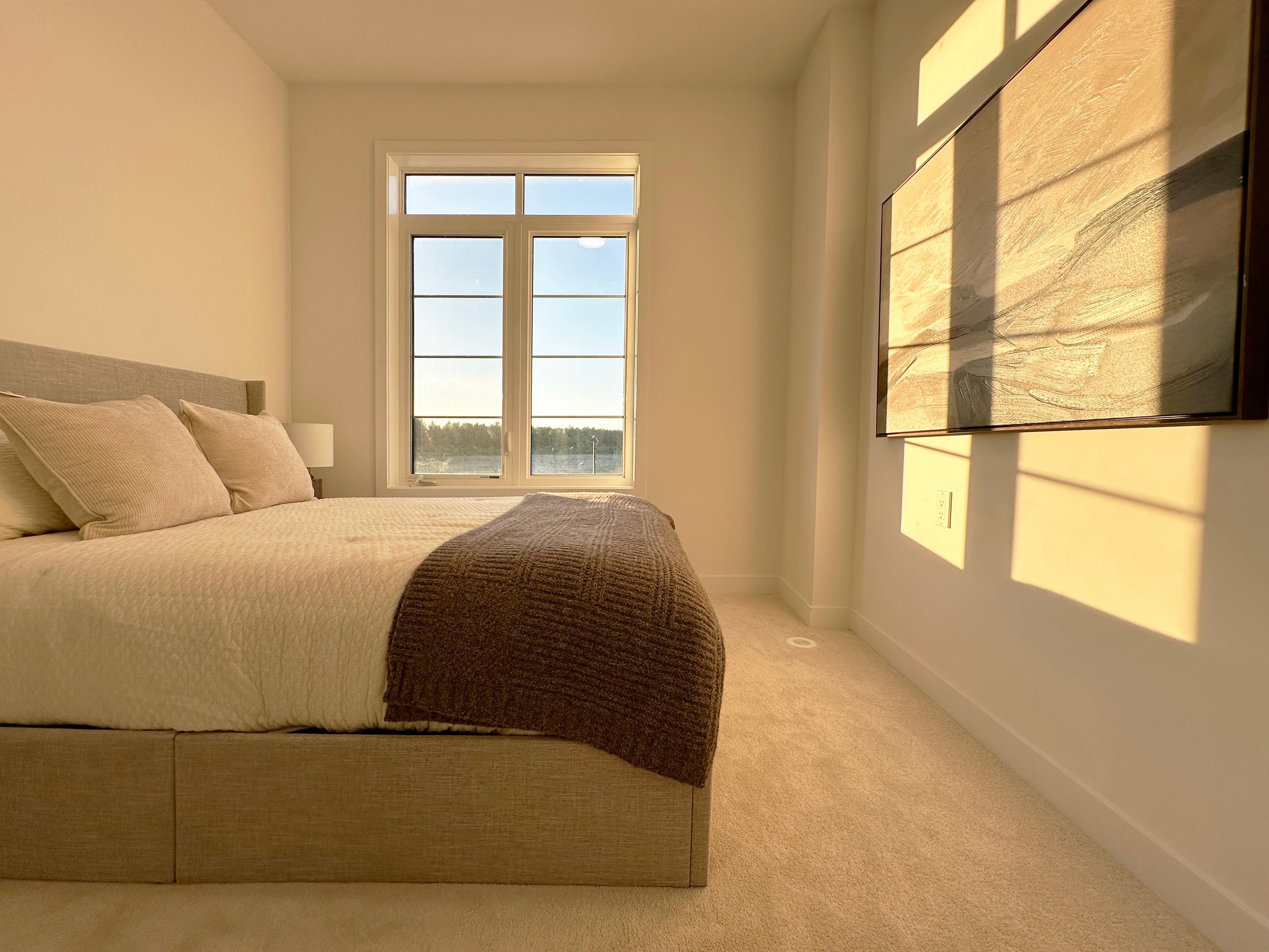 A cozy bedroom with beige walls, a bed with cream-colored bedding and pillows, a window with sunlight streaming in, and a large abstract painting on the wall.